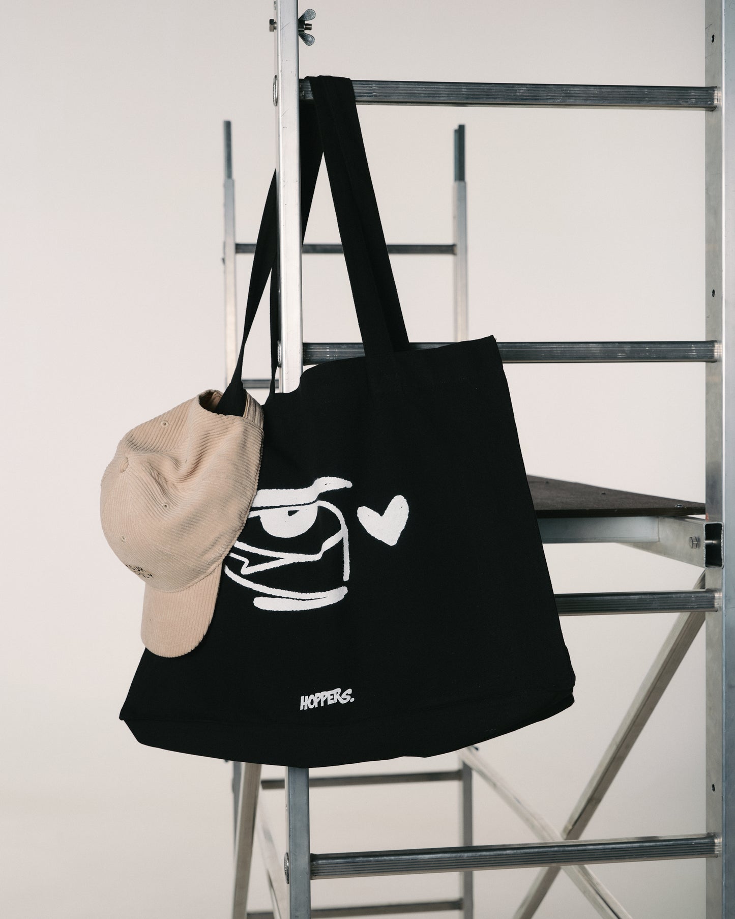 A black tote bag with a white cartoon face and heart design hangs on a metal ladder. A beige cap is draped over the bag's handle. Minimalist setting.