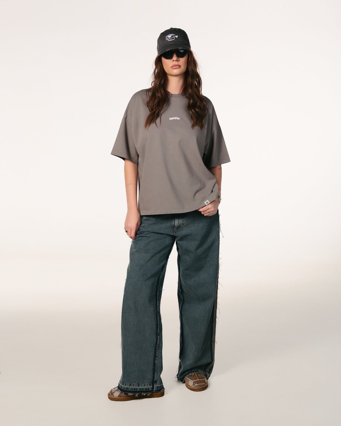 Woman with long brown hair, wearing sunglasses, a dark cap, gray oversized Hoppers Hip Hop Takeovers T-shirt, and blue jeans. She stands relaxed against a plain background.