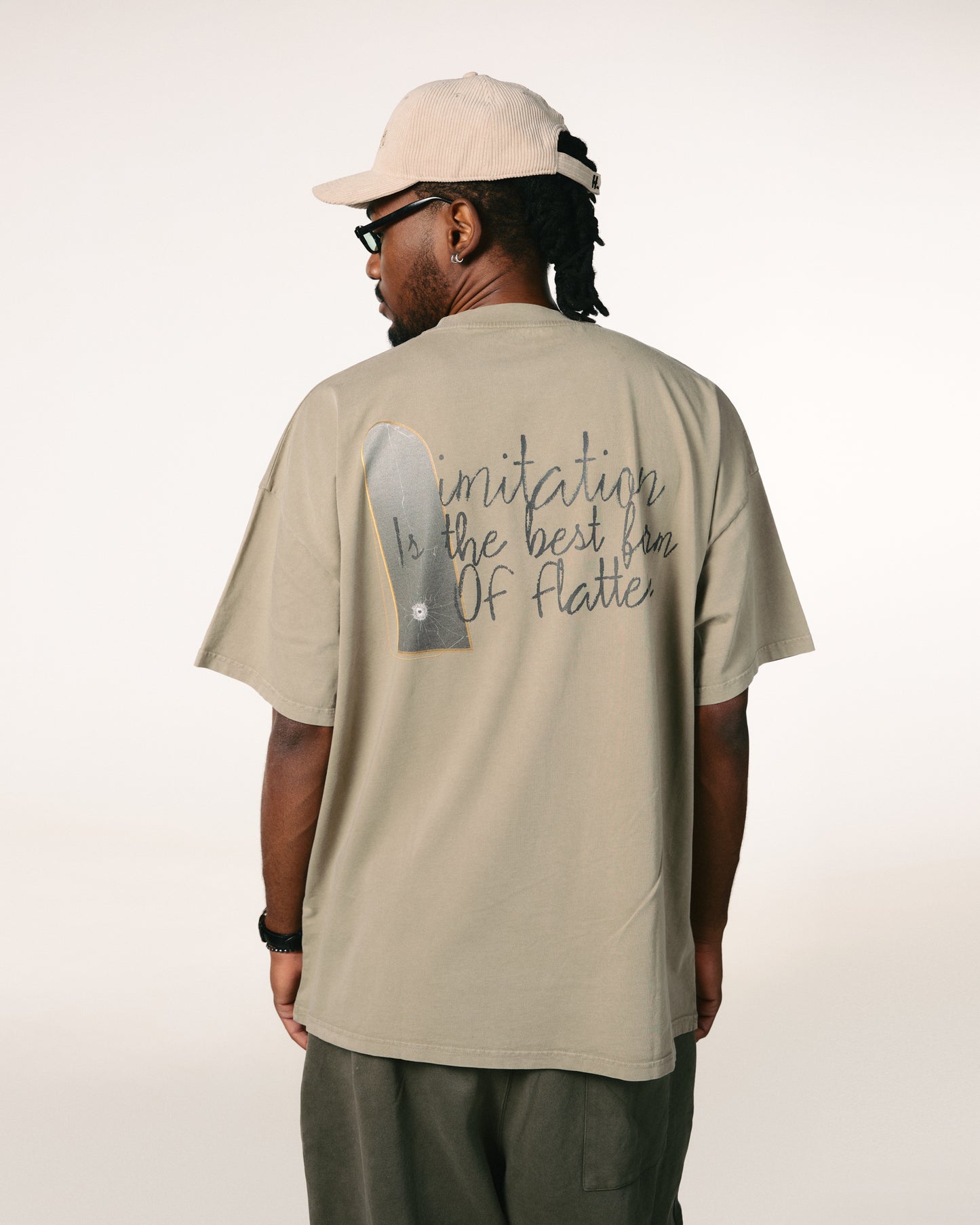 A man in a beige cap and glasses is seen from behind, wearing a tan shirt with the text "Imitation is the best form of flattery." The setting is minimal and neutral.