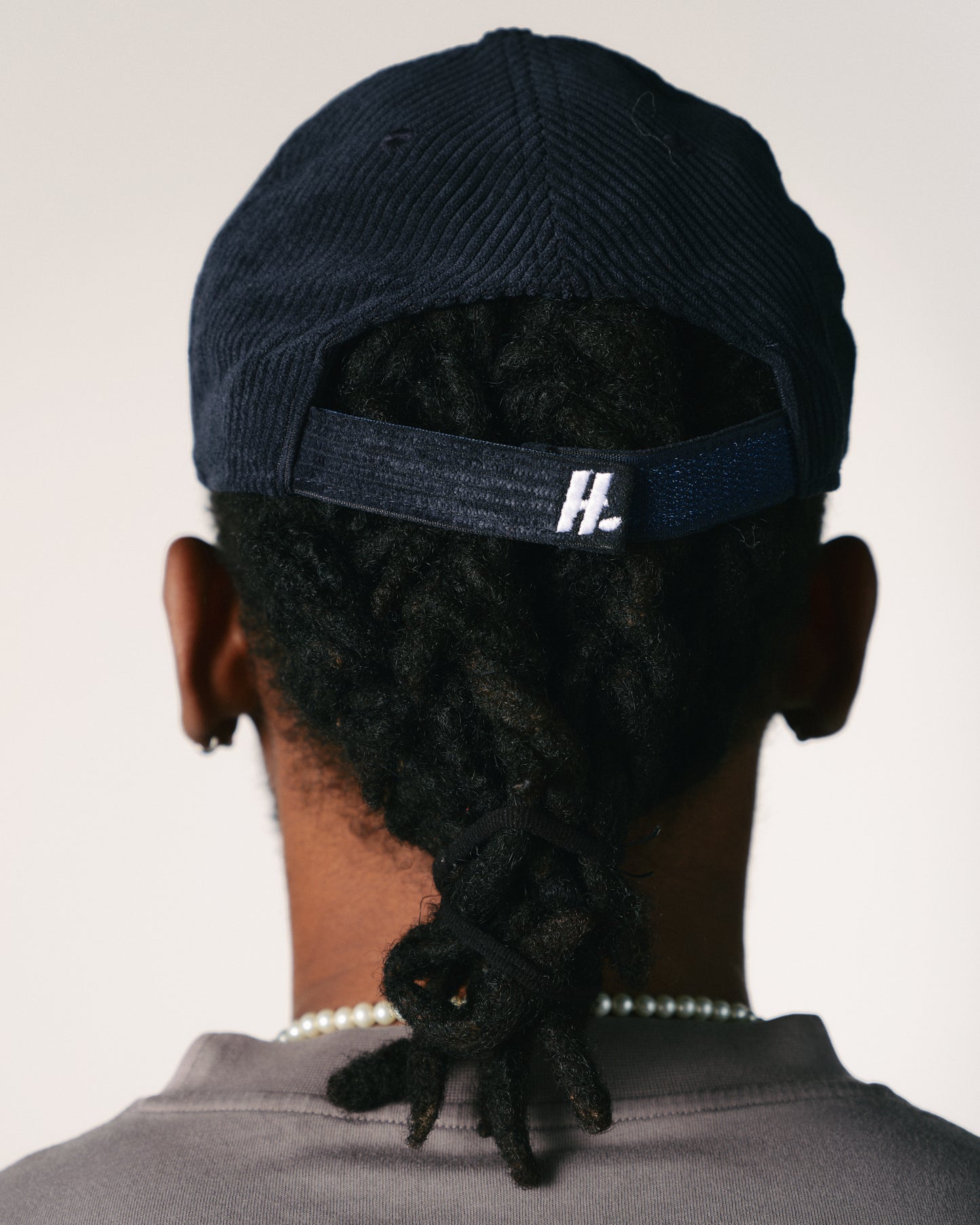 The back view of a person wearing a navy blue cap with dreadlocks tied back. A pearl necklace is visible on the neck against a neutral background.
