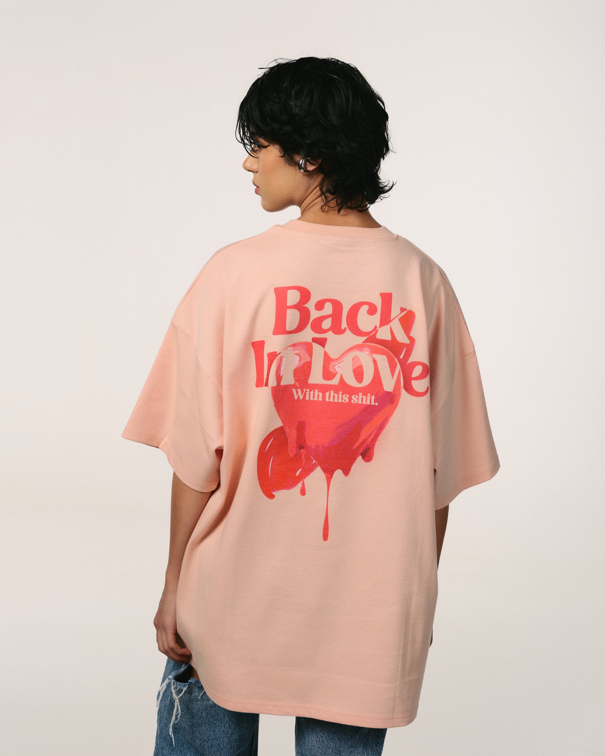 A person with  t short Hoppers, hip-hop takeovers , dark hair is wearing a peach pink  colored T-shirt with bold red text and a heart design on the back, reading "Back In Love." 100% organic cotton, eco friendly, premium quality t shirt