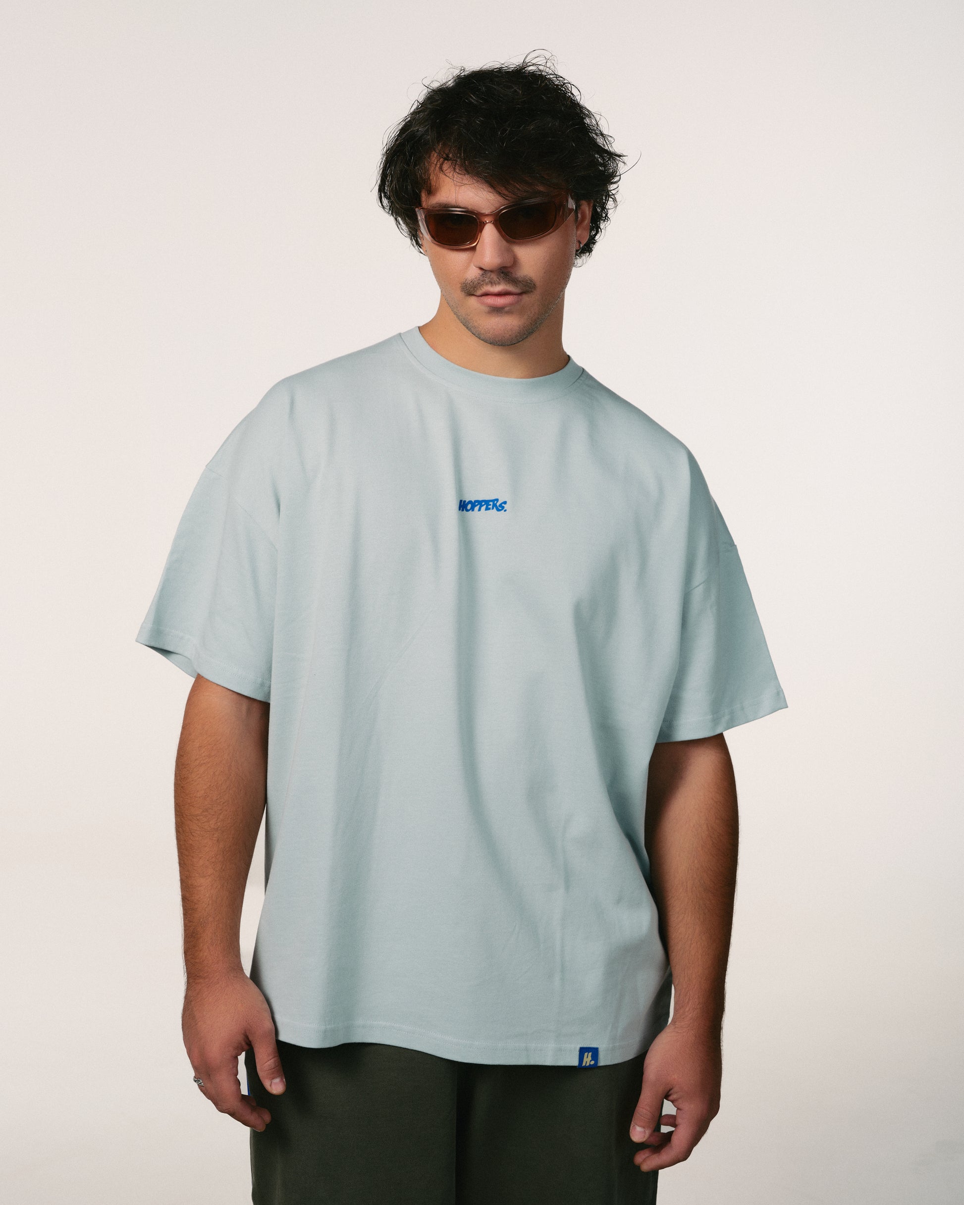 Man with tousled hair in sunglasses wears a loose, oversize, light gray-blue baby blue T-shirt and dark pants, exuding a relaxed, casual vibe against a plain backdrop. Hoppers hip hop takeovers logo, 100% organic cotton bumbac