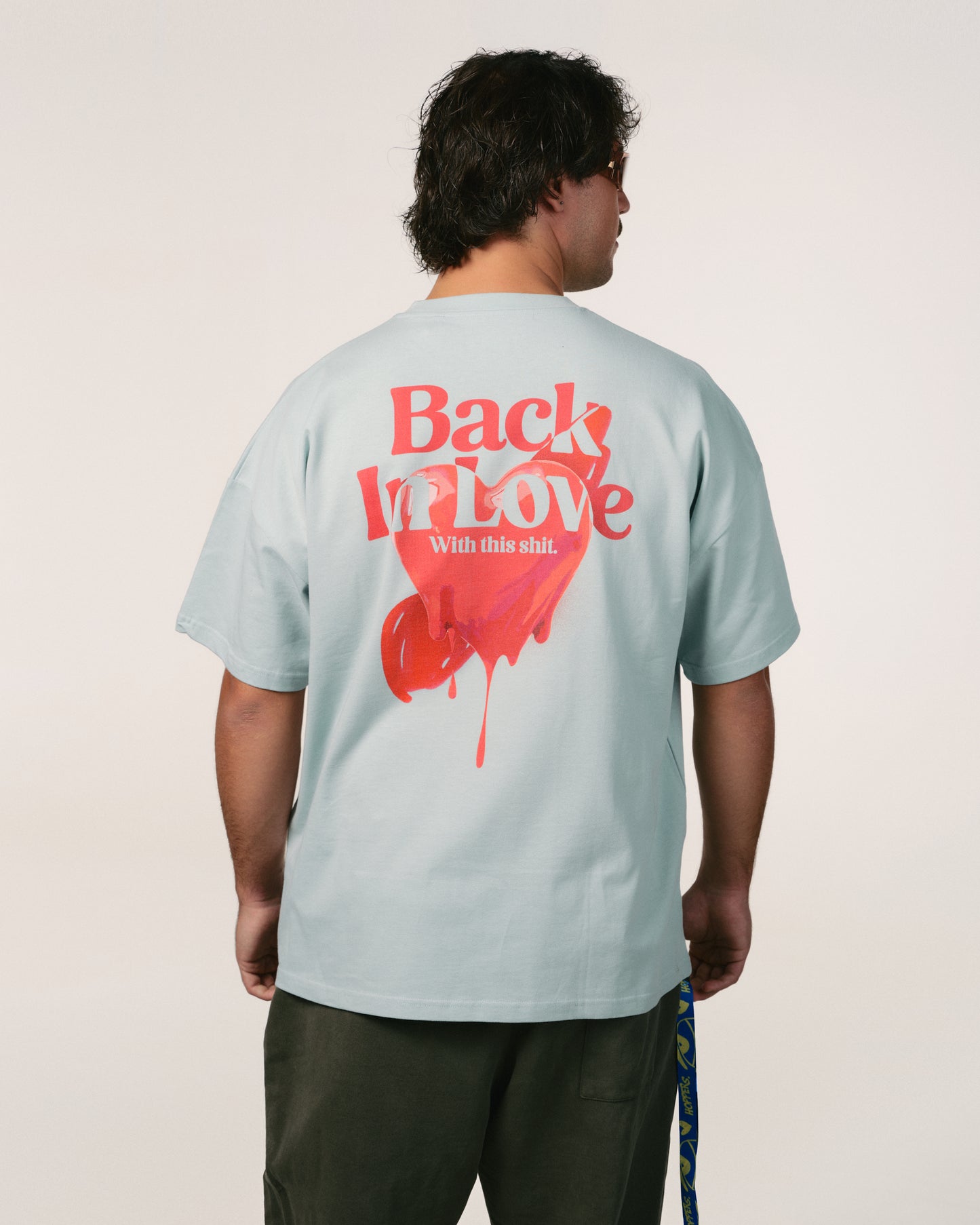 Man facing away, wearing a light blue, baby blue 100% organic cotton bumbac T-shirt Hoppers Hip hop takeovers with red text and a heart graphic saying "Back In Love." Casual tone, relaxed vibe.