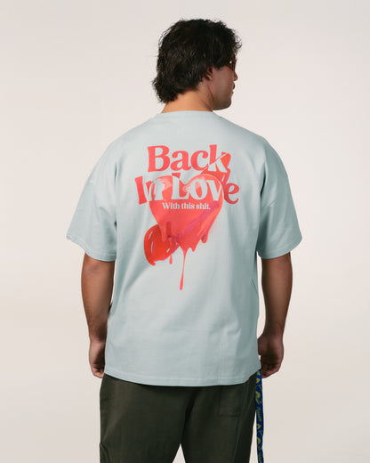 Man facing away, wearing a light blue, baby blue 100% organic cotton bumbac T-shirt Hoppers Hip hop takeovers with red text and a heart graphic saying "Back In Love." Casual tone, relaxed vibe.