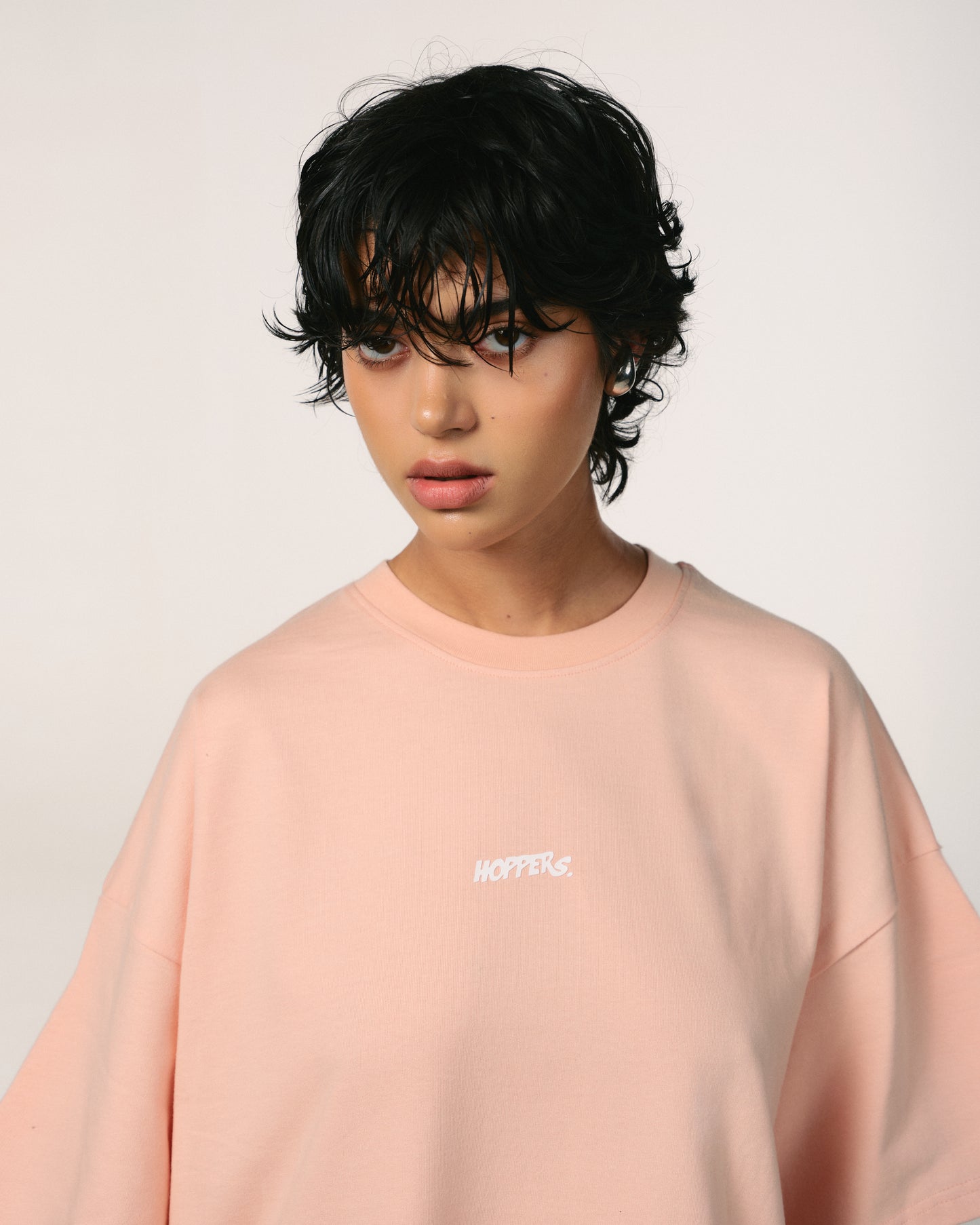 A person with short, wet-look black hair wears a peach pink oversized 100% organic cotton premium t shirt with "HOPPERS" " Back in love" or Hip-Hop Takeovers  logo in white. The backdrop is plain, creating a serene mood.