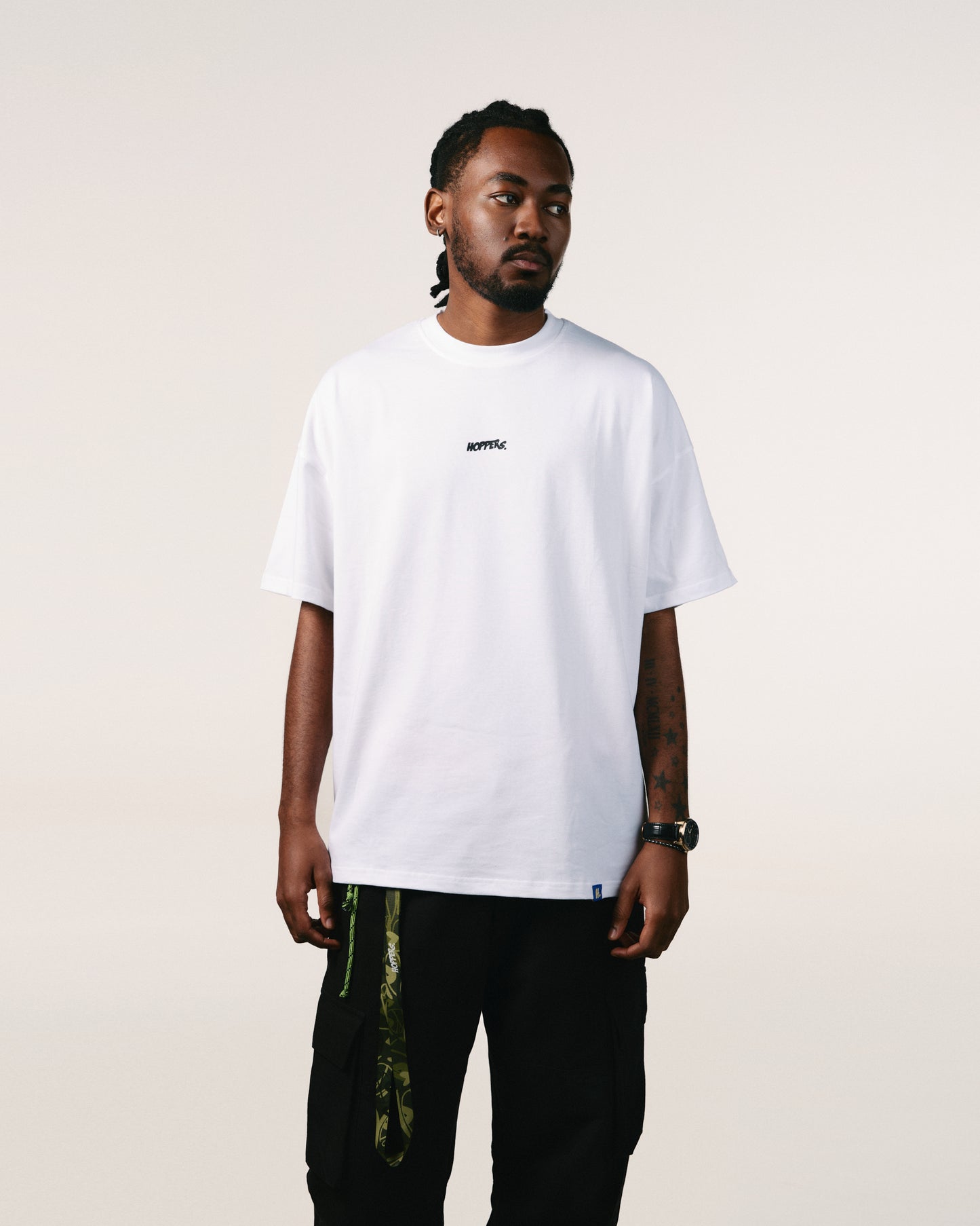 A person with braided hair stands against a neutral background wearing a white "Hoppers" Hip-Hop takeovers T-shirt and black cargo pants, exuding a relaxed, casual vibe, streetwear.