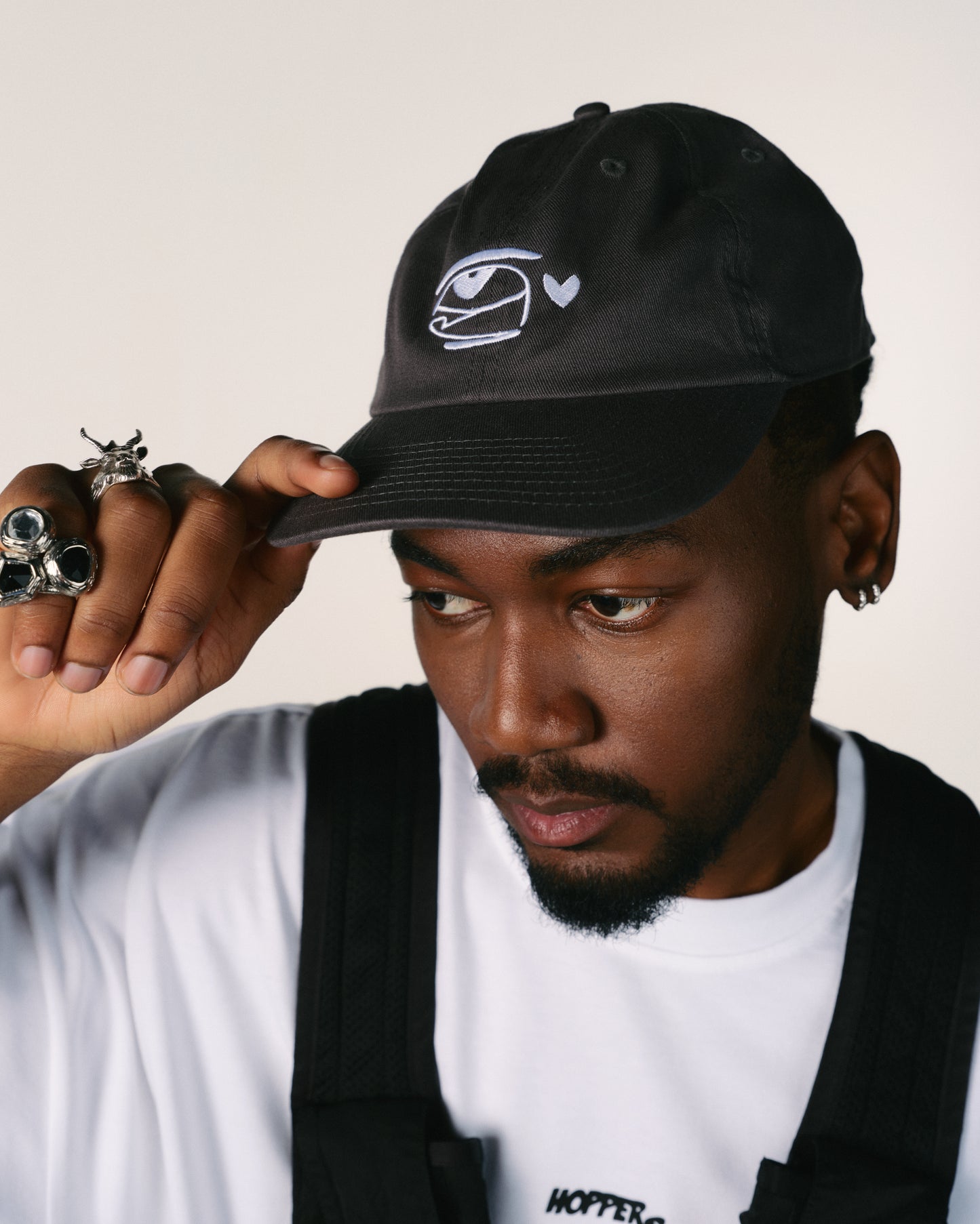 Man wearing a black cap with a hoppers hip hop takeovers logo, holding it with one hand.
