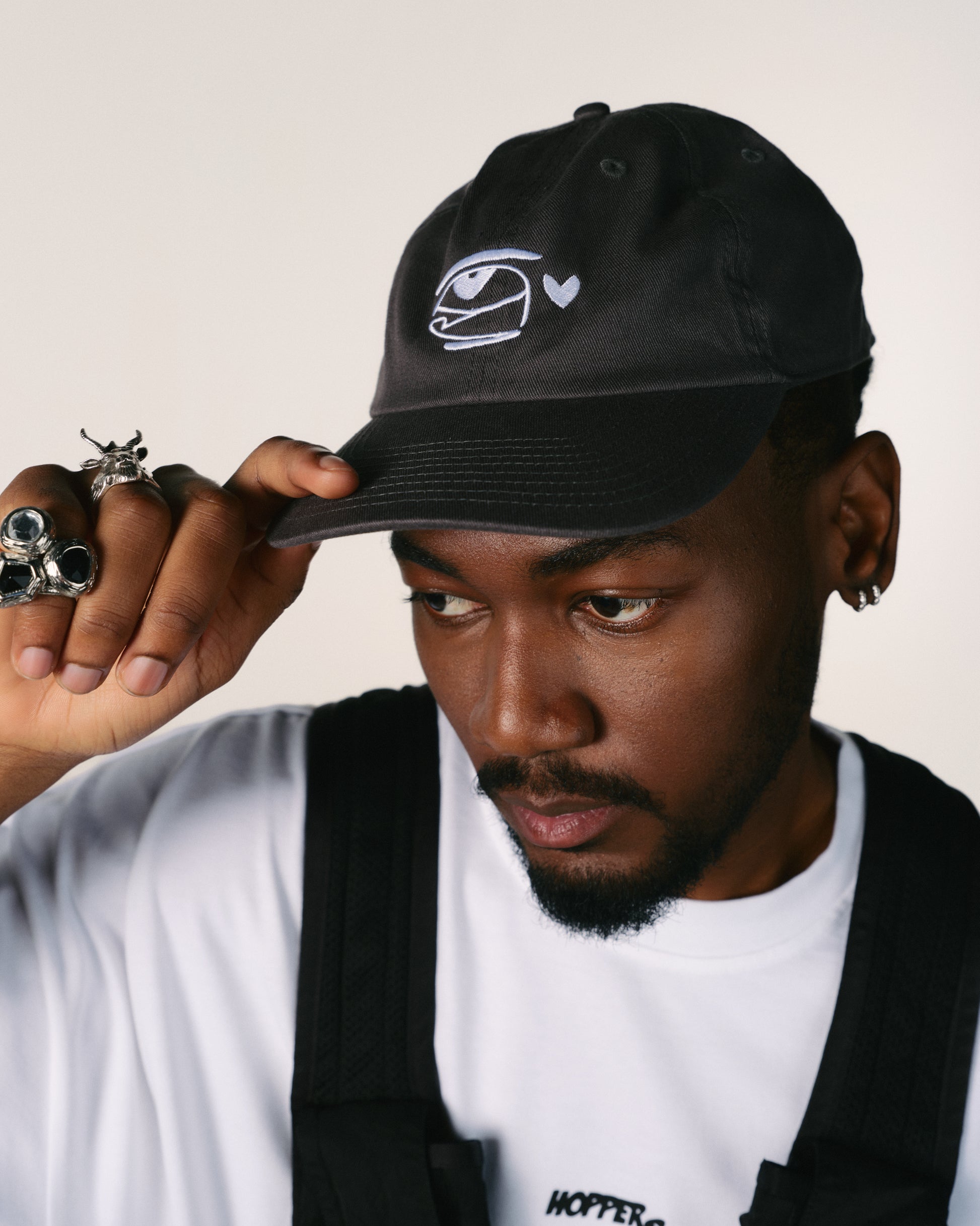 Man wearing a black cap with a hoppers hip hop takeovers logo, holding it with one hand.