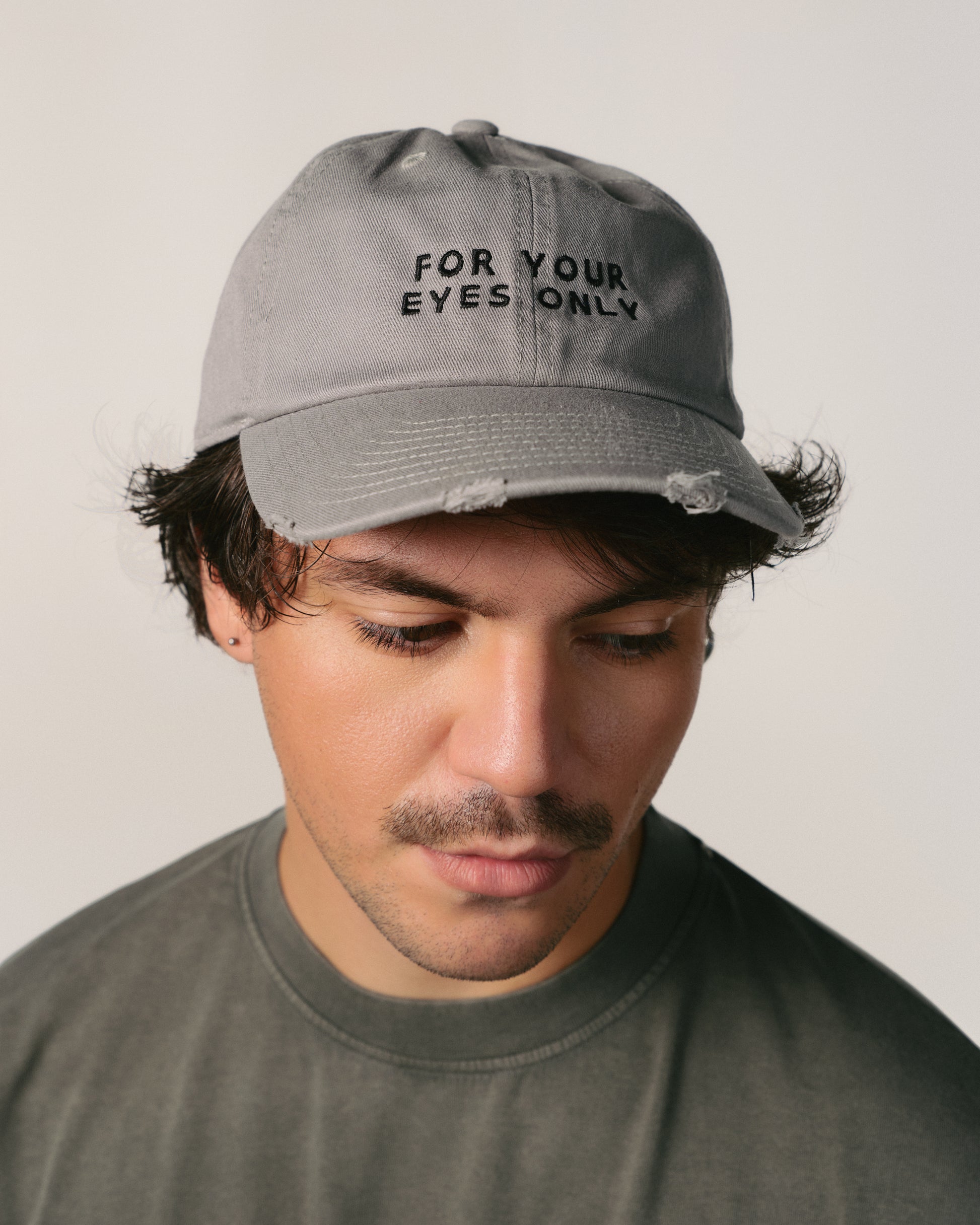A person wearing a gray cap with "For Your Eyes Only" text. They look downward with a neutral expression, wearing a matching gray shirt.