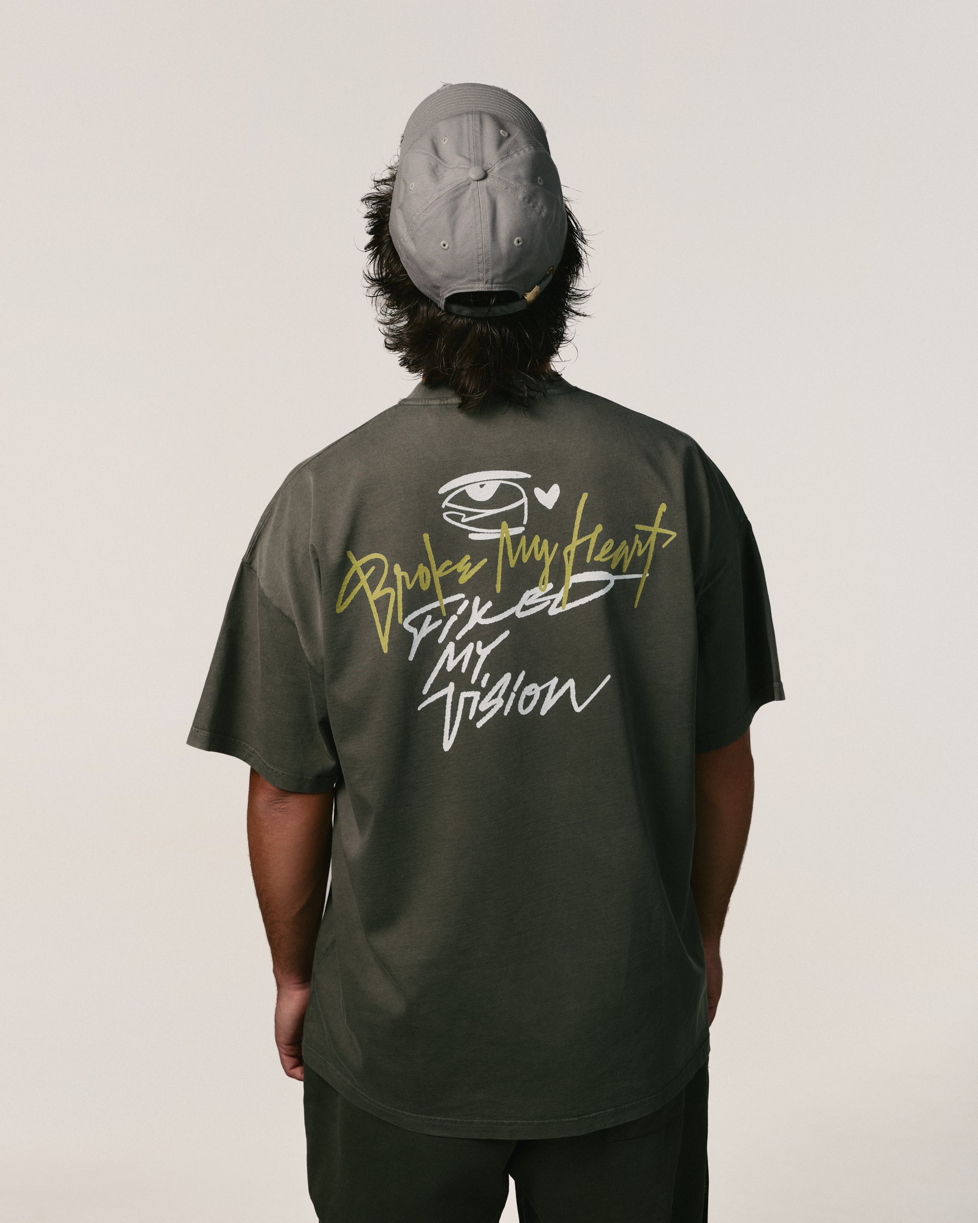 Back view of a person in a gray cap and dark shirt with text: "Broke My Heart Fixed My Vision." The style is artistic and conveys resilience.