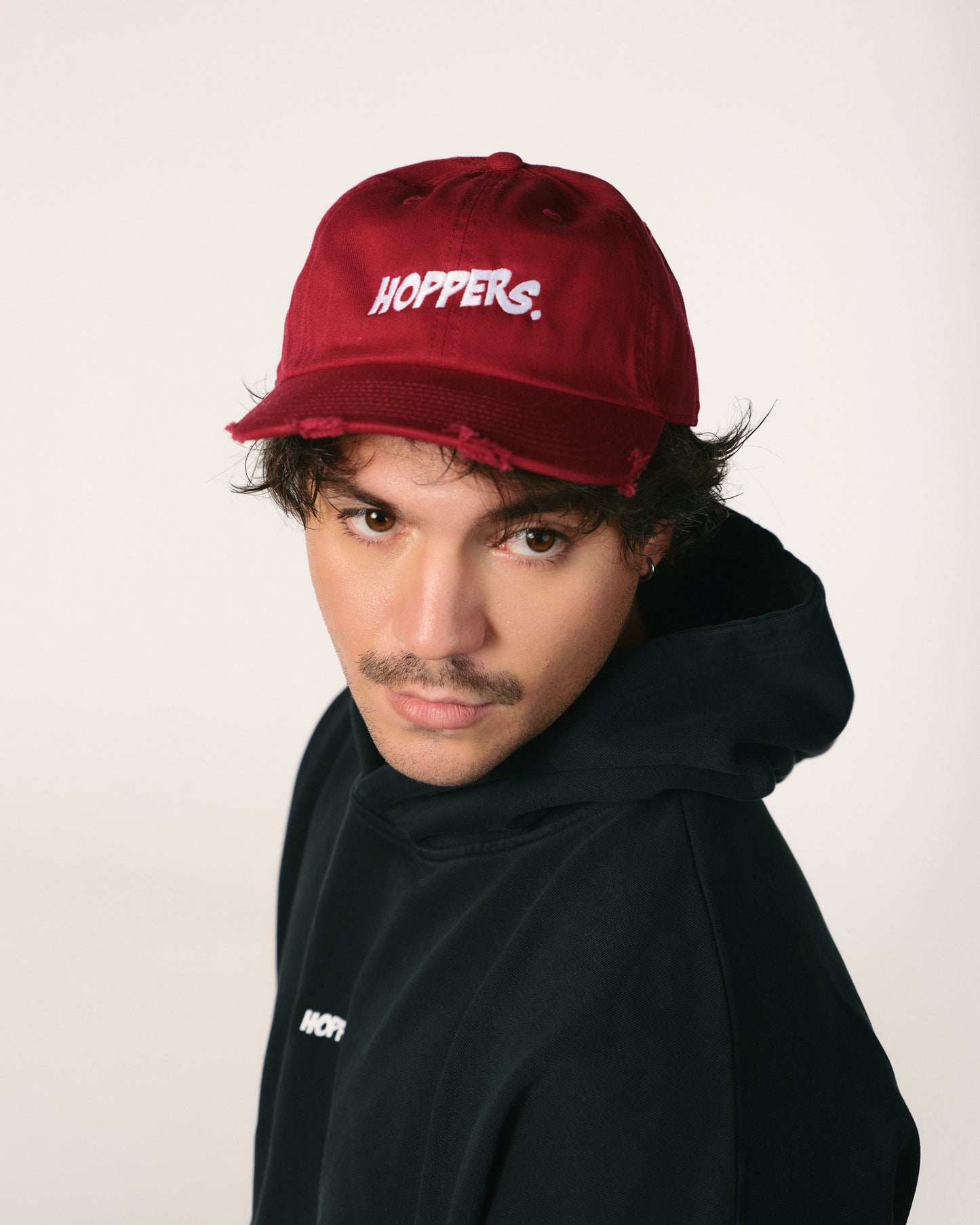 Young man in a red cap and black hoodie gazes directly at the camera with a serious expression. The cap features the word "HOPPERS" in white text.