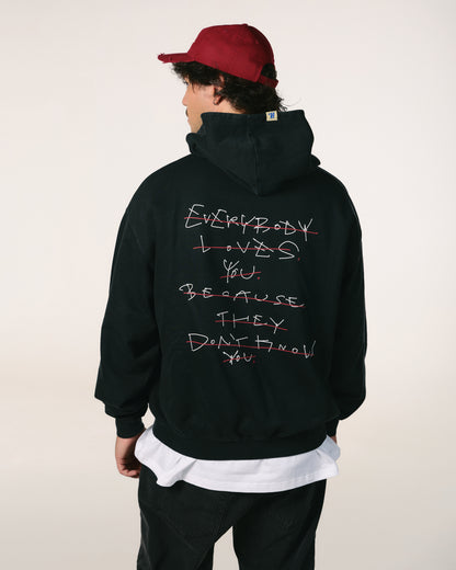 A person wearing a red cap and a black hoodie with white text reading, "Everybody loves you until they don't know you," crossed out in red. The mood is introspective.
