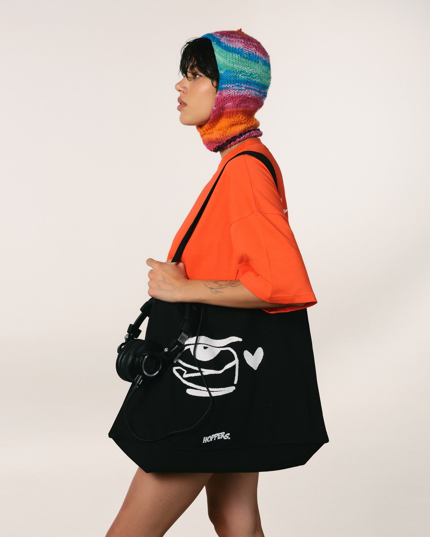 A person in profile wears a bright orange Hoppers t shirt and a colorful knit balaclava, holding a large black hoppers fashion bag with a white "for your eyes only" logo and heart design. The tone is modern and stylish.