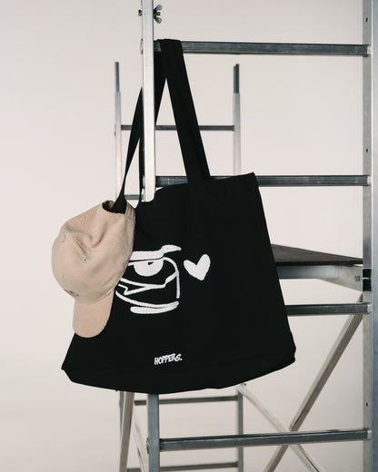 A black tote bag with a white cartoon face and heart design hangs on a metal ladder. A beige cap is draped over the bag's handle. Minimalist setting.