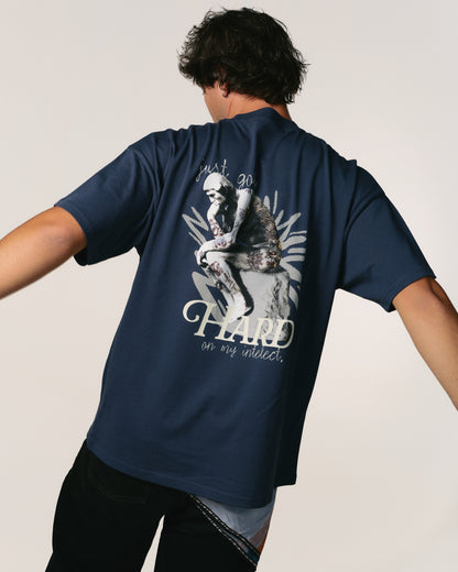 A person facing away, wearing a navy blue  Hoppers Hip Hop Takeovers T-shirt with a graphic of a thinker statue and the text "just go HARD on my intellect," against a plain background.