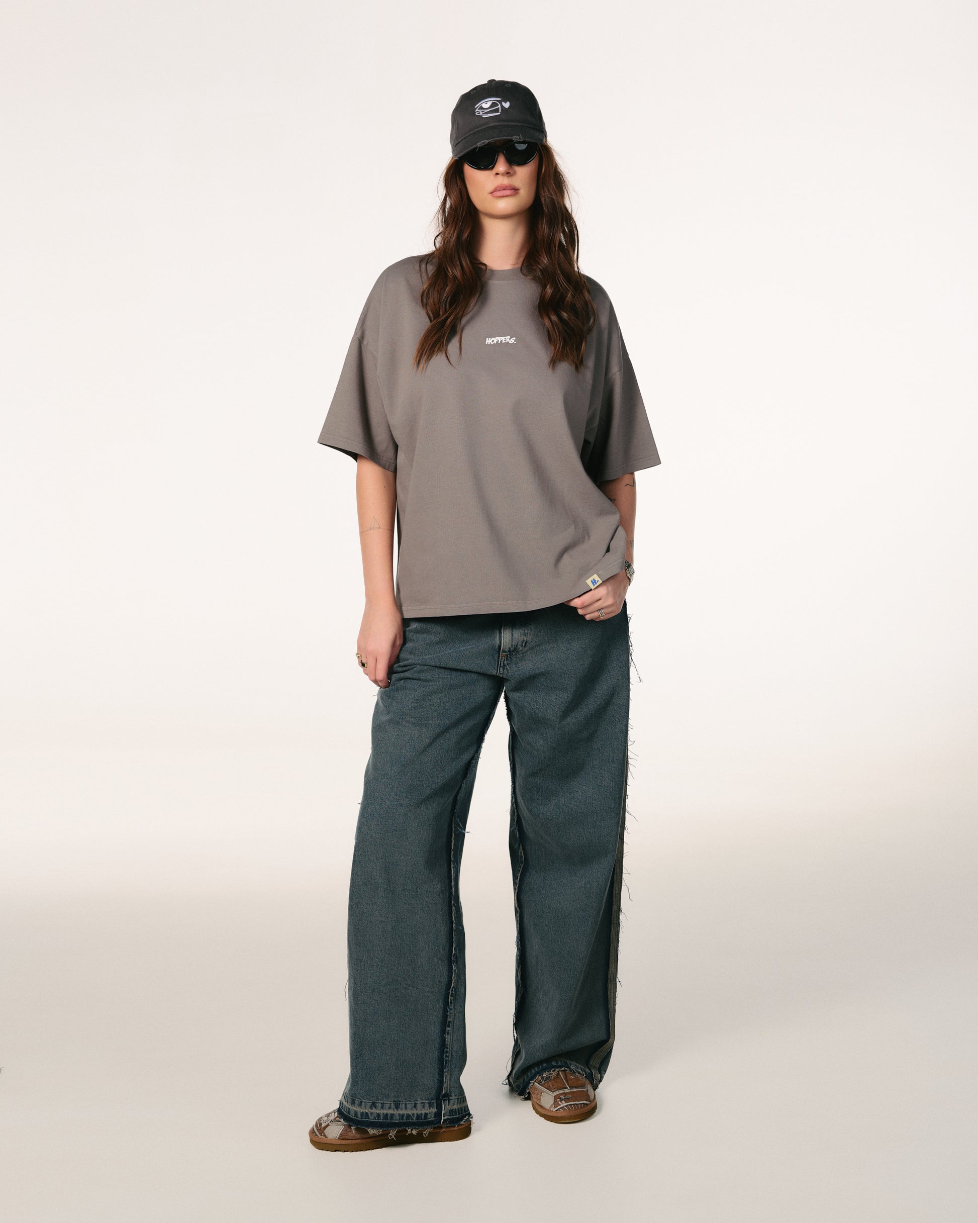Woman with long brown hair, wearing sunglasses, a dark cap, gray oversized Hoppers Hip Hop Takeovers T-shirt, and blue jeans. She stands relaxed against a plain background.