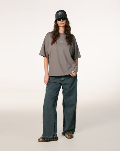 Woman with long brown hair, wearing sunglasses, a dark cap, gray oversized Hoppers Hip Hop Takeovers T-shirt, and blue jeans. She stands relaxed against a plain background.