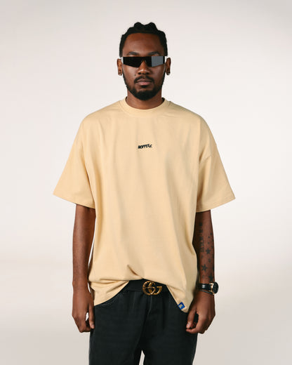 Man in a beige T-shirt and black jeans stands confidently. He wears dark sunglasses and has a watch on his left wrist. Neutral background.