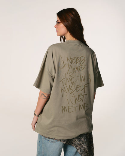 A person with long hair and glasses faces away, wearing an oversized olive green  Hoppers Hip Hop Takeovers T-shirt with bold text: "I need some time with myself I just met me." Relaxed, introspective mood.