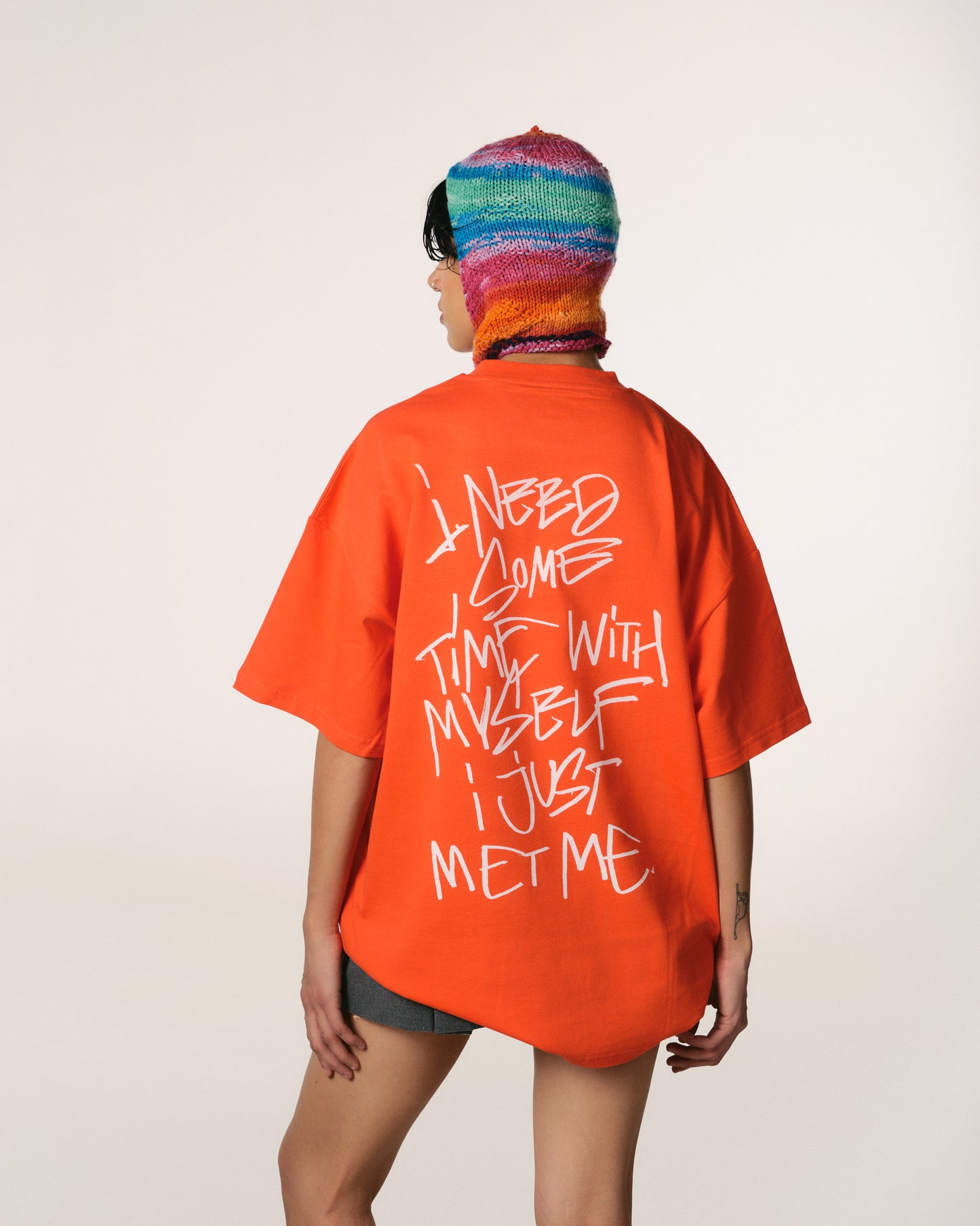 A person wearing an orange Hoppers Hip Hop Takeovers T-shirt with graffiti-style white text reading, "I need some time with myself, I just met me." They also wear a colourful knit hat.
