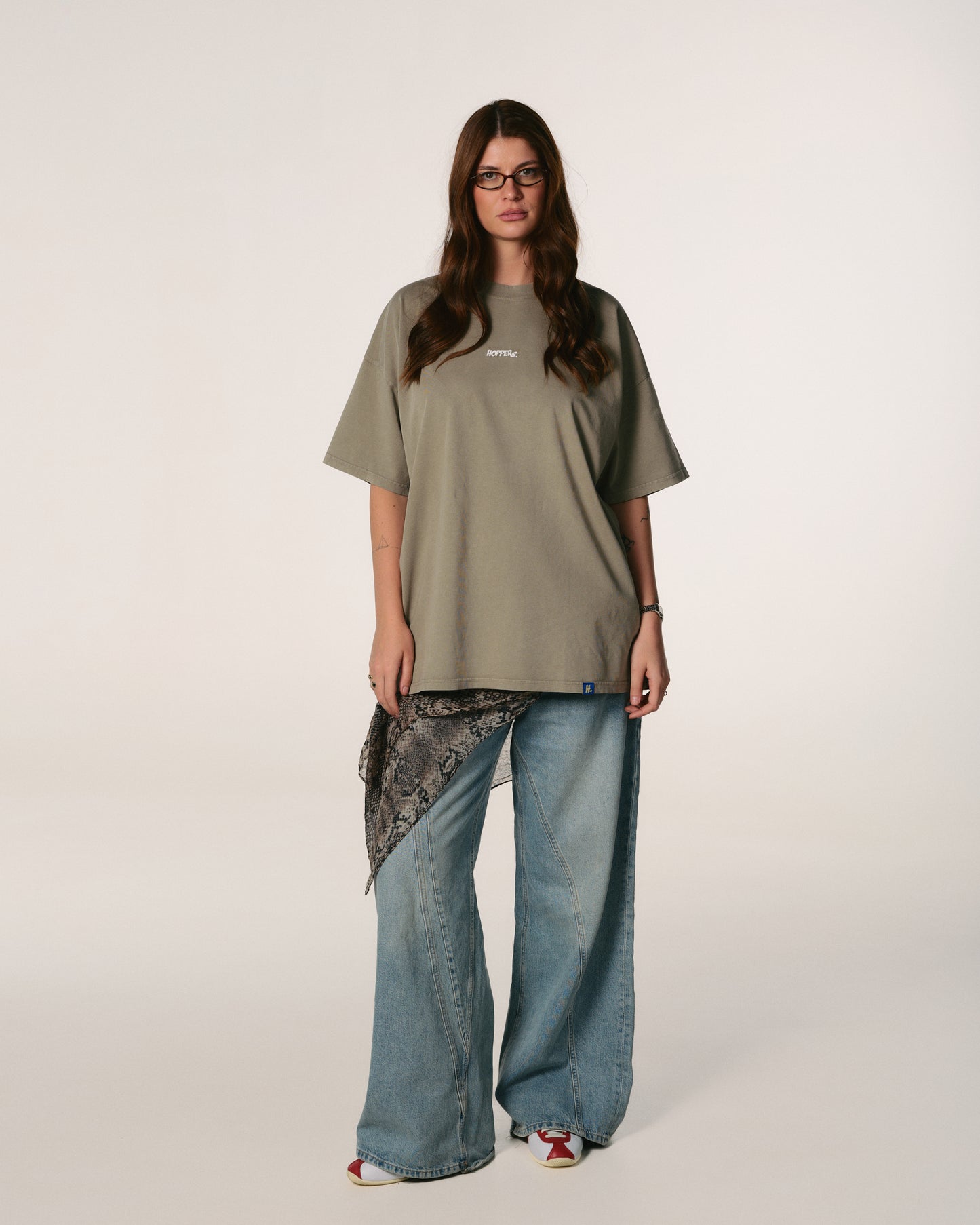 A woman with long hair wearing glasses stands confidently. She is dressed in a loose, olive-green Hoppers Hip Hop takeovers T-shirt with text, paired with jeans and a patterned scarf.