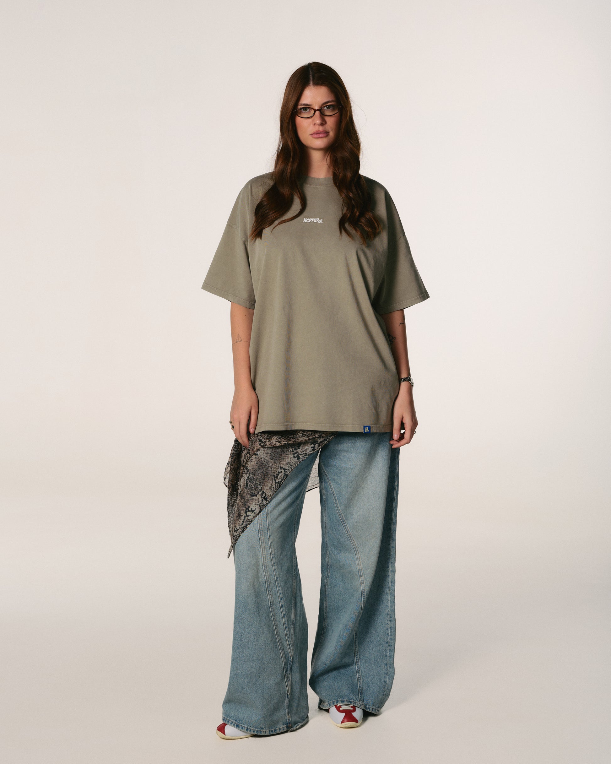 A woman with long hair wearing glasses stands confidently. She is dressed in a loose, olive-green Hoppers Hip Hop takeovers T-shirt with text, paired with jeans and a patterned scarf.