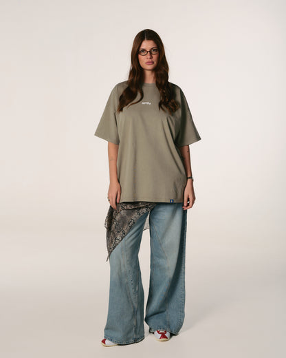 A woman with long hair wearing glasses stands confidently. She is dressed in a loose, olive-green Hoppers Hip Hop takeovers T-shirt with text, paired with jeans and a patterned scarf.