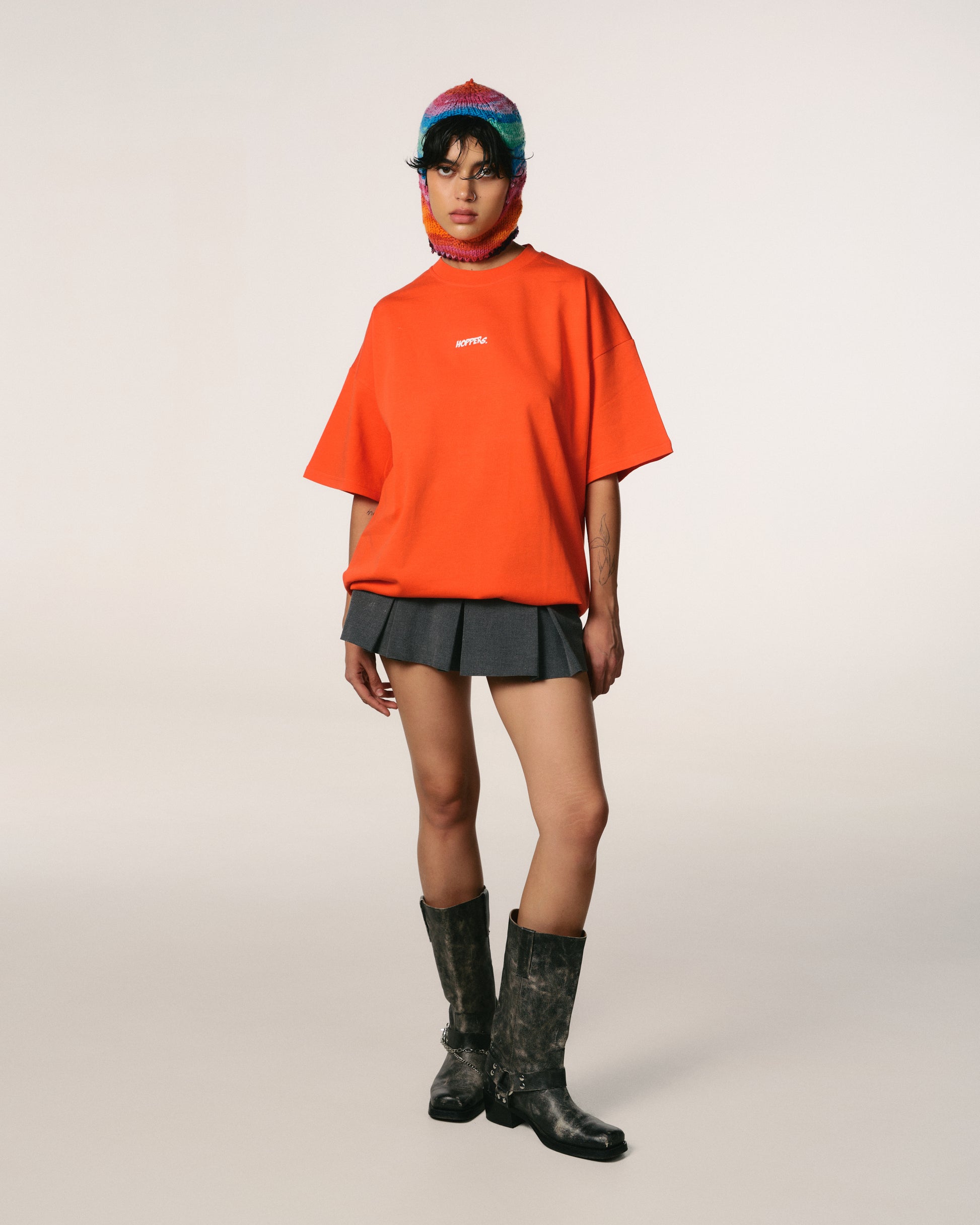 A model stands wearing a loose orange Hoppers  shirt, grey pleated skirt, and black boots. A colourful  knit balaclava adds a playful touch. The setting is minimal, creating a bold, stylish vibe.