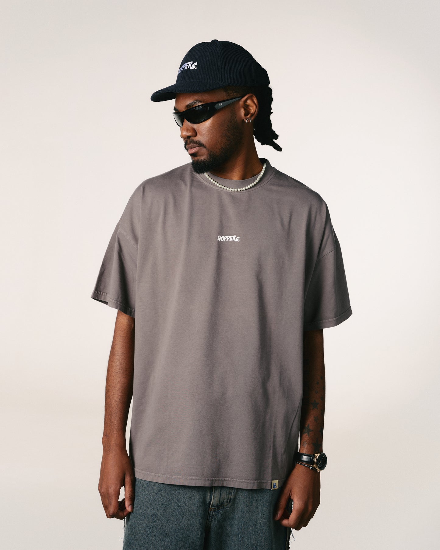 Man in a gray  Hoppers Hip Hop Takeovers T-shirt and navy cap with sunglasses, looking left. He wears a necklace and watch, conveying a casual and cool vibe.