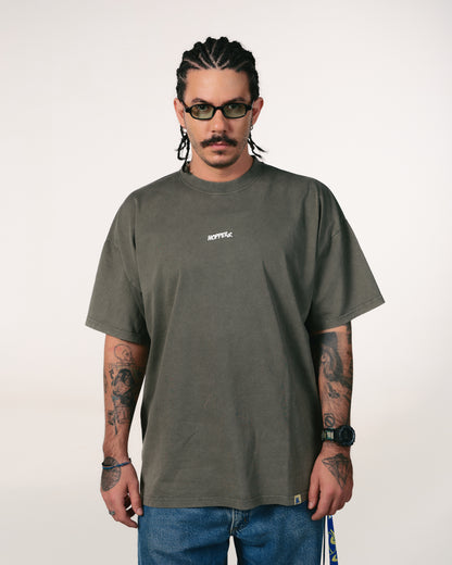 Man with cornrows and glasses in a gray oversized T-shirt and blue jeans. He has tattoos on his arms and a serious expression. White background.