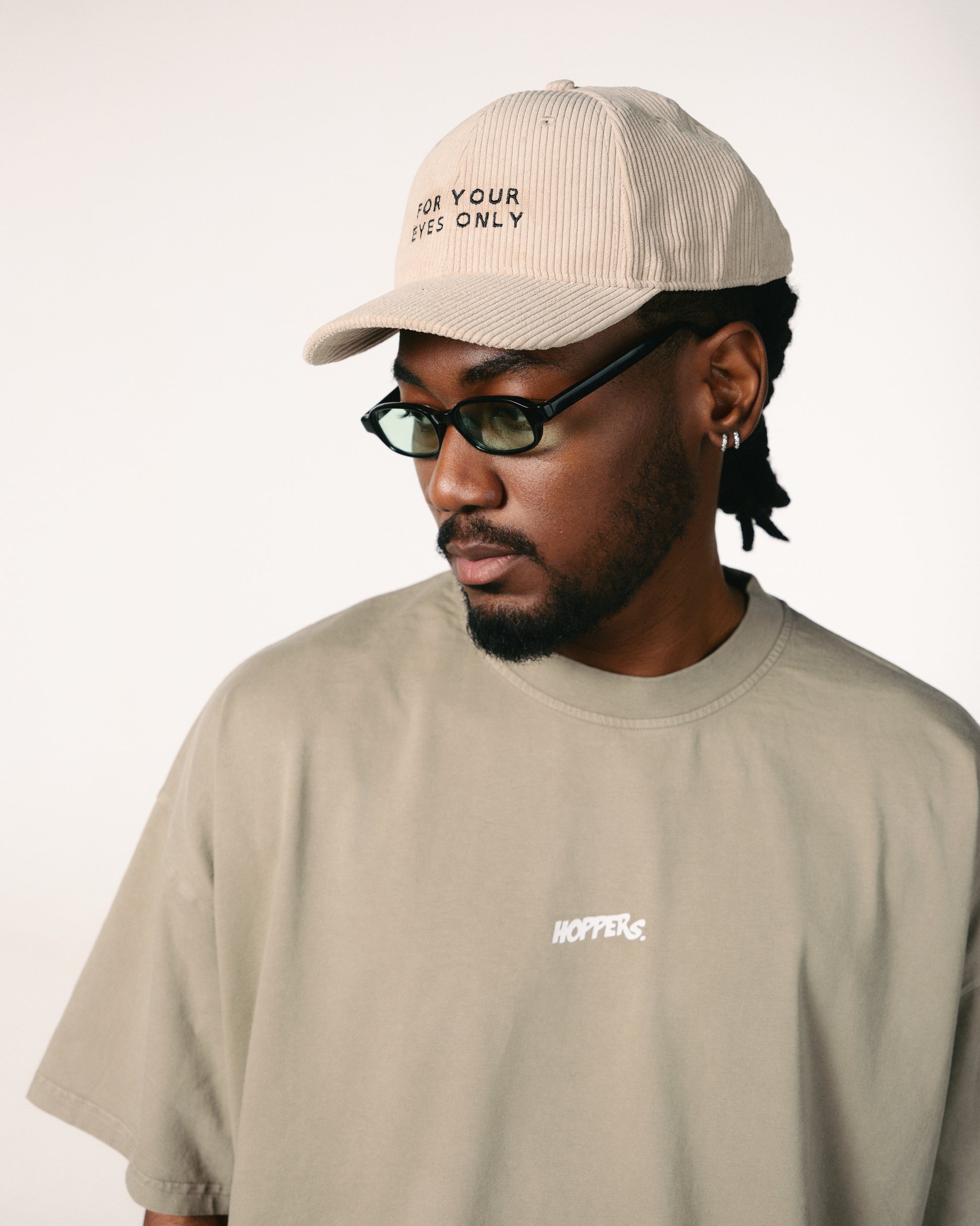 Man wearing olive shirt and beige baseball cap embroidered with "For Your Eyes Only." He sports black glasses, exuding a calm, thoughtful vibe.