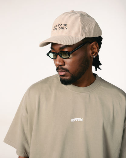 Man wearing olive shirt and beige baseball cap embroidered with "For Your Eyes Only." He sports black glasses, exuding a calm, thoughtful vibe.