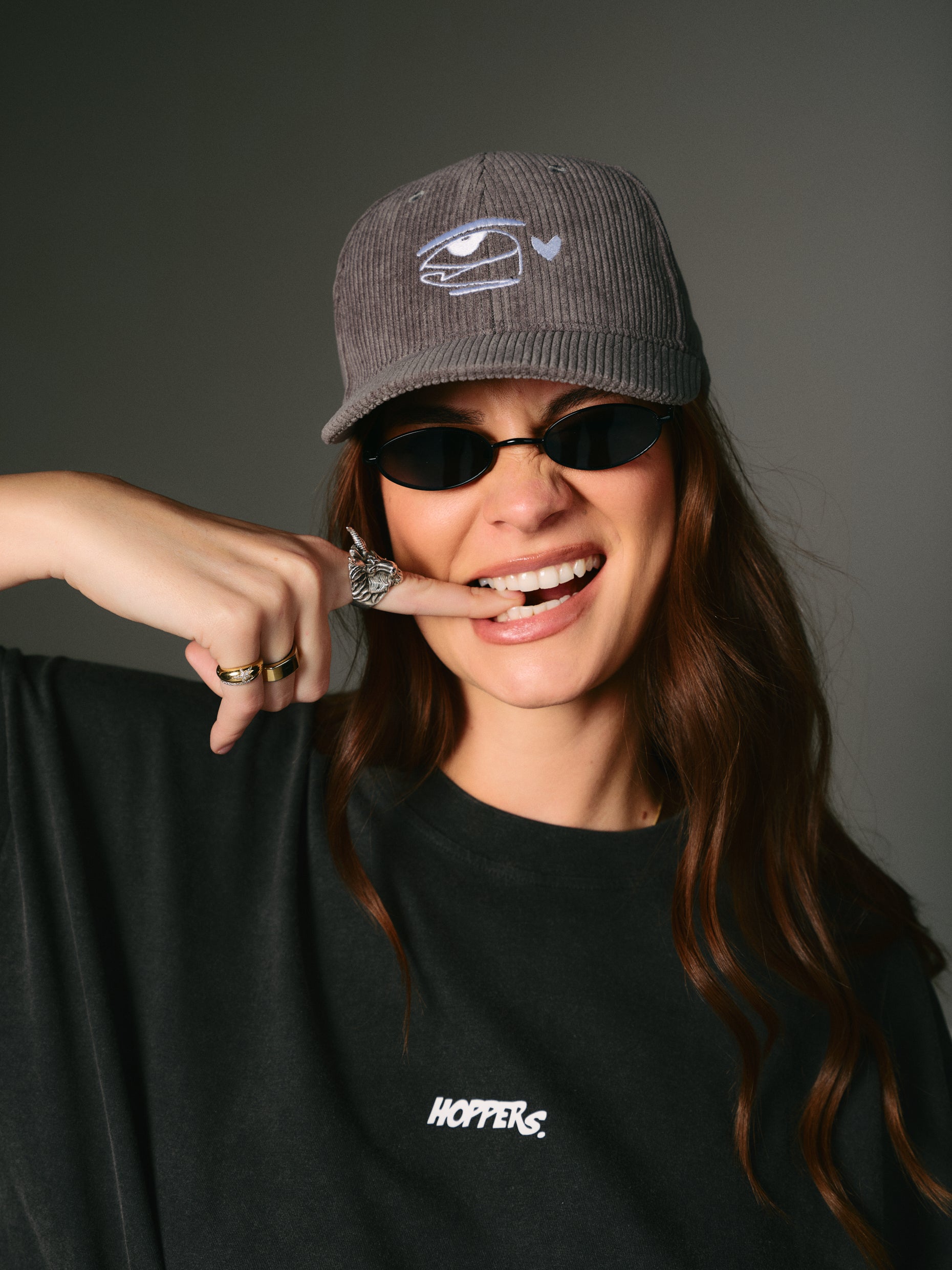 A woman playfully bites her finger, wearing a grey  Corduroy cap with an eye logo, black sunglasses, and a dark "Hoppers" T-shirt. She exudes a carefree vibe.
