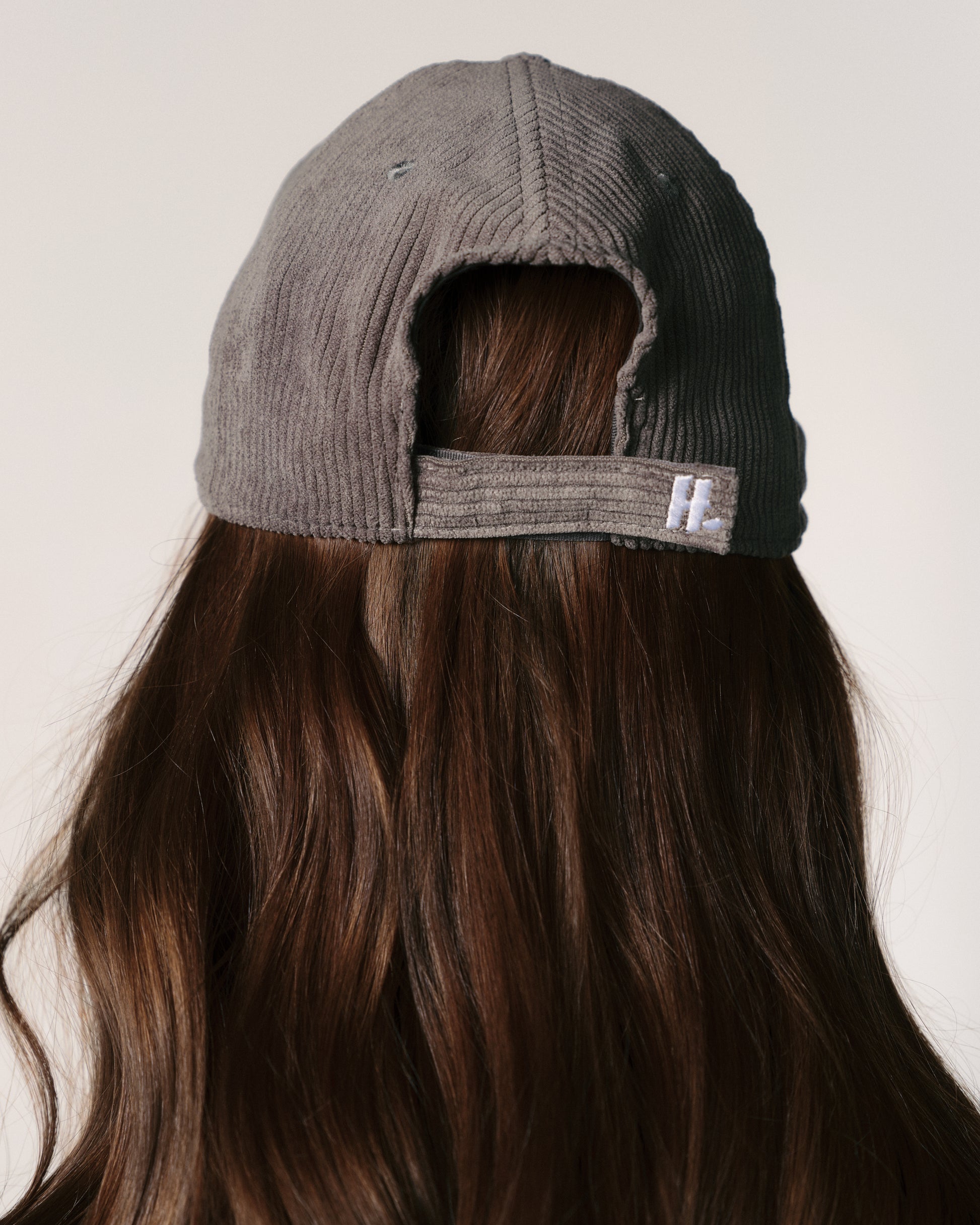 A person with long brown hair wears a gray corduroy baseball cap backwards. The background is neutral, emphasizing the casual and relaxed tone.