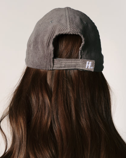 A person with long brown hair wears a gray corduroy baseball cap backwards. The background is neutral, emphasizing the casual and relaxed tone.