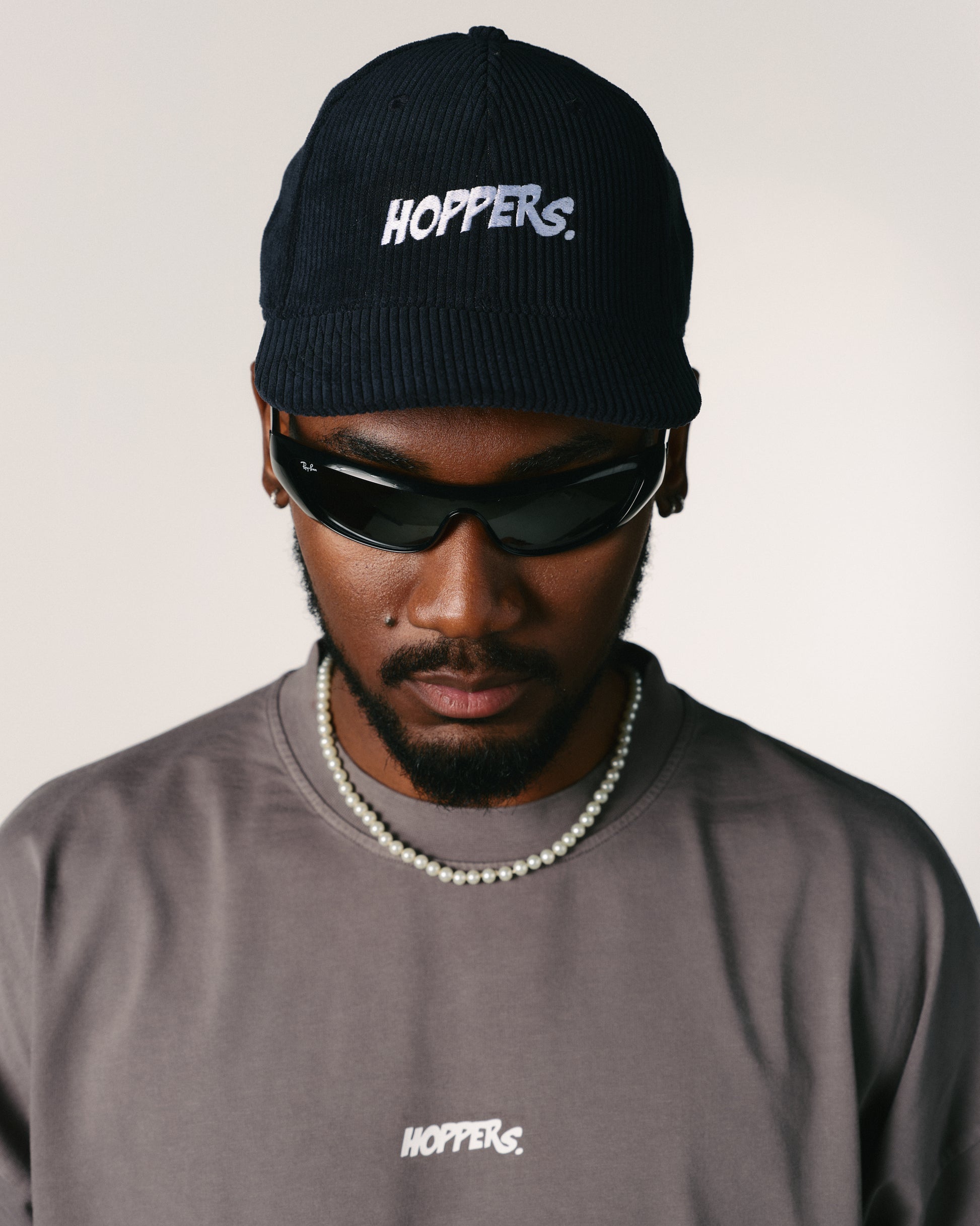 A person wearing a black cap and sunglasses looks down, exuding a cool demeanor. The cap and shirt both display the "Hoppers" logo in white.