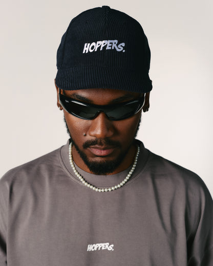 A person wearing a black cap and sunglasses looks down, exuding a cool demeanor. The cap and shirt both display the "Hoppers" logo in white.