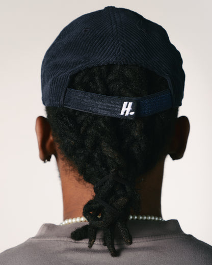 The back view of a person wearing a navy blue cap with dreadlocks tied back. A pearl necklace is visible on the neck against a neutral background.