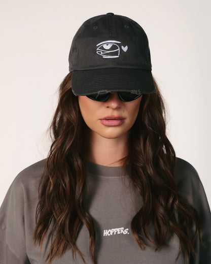 A woman with wavy brown hair wears a black cap with an abstract eye and heart design. She has sunglasses and a gray shirt labeled "HOPPERS." Sporting a neutral expression.