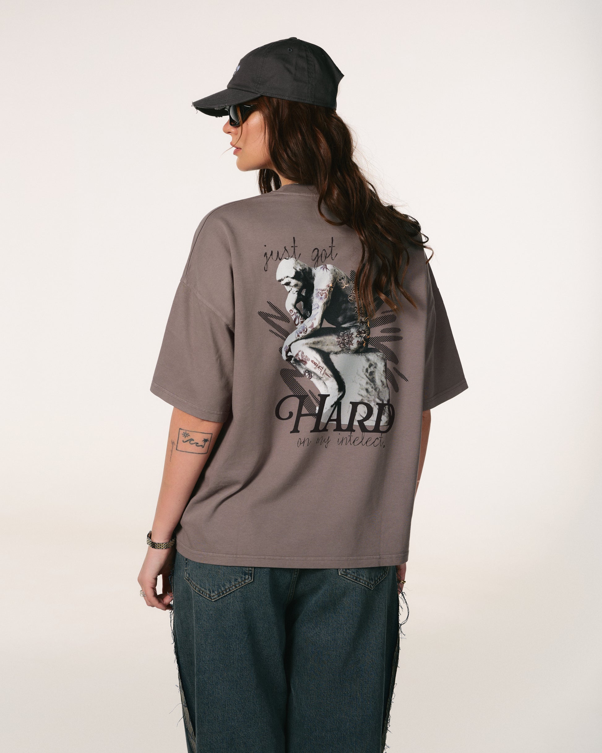 A person wearing a gray cap, sunglasses, and a loose gray T-shirt with an artistic design and text on the back. They have wavy hair and casual jeans.