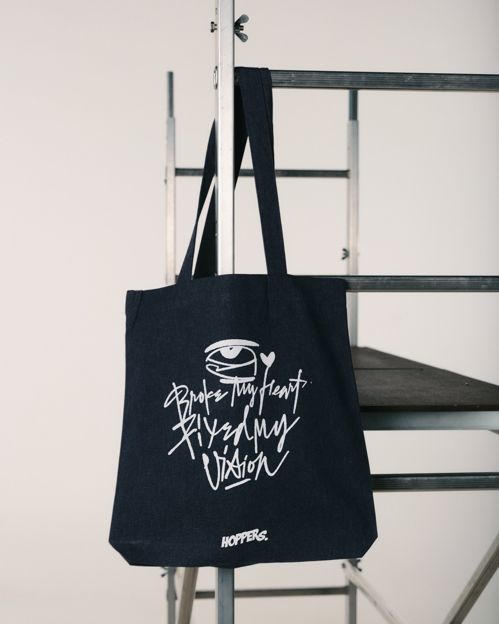 Black tote bag with white text and logo on a metal chair against a white wall
