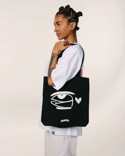 Person holding a black tote bag with a white logo and text on a plain background