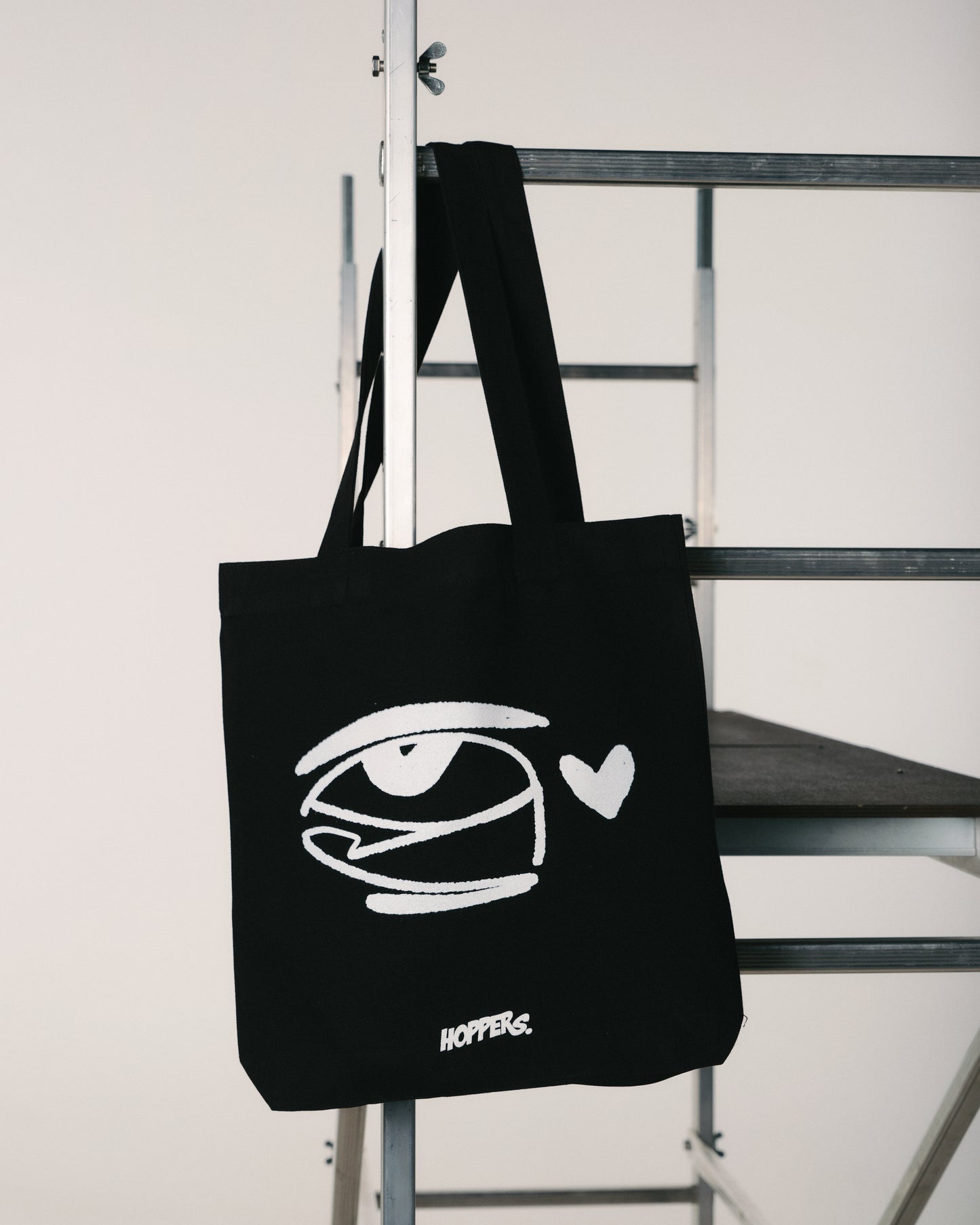 Black tote bag with white eye design and 'Hopper' branding on a metal staircase.