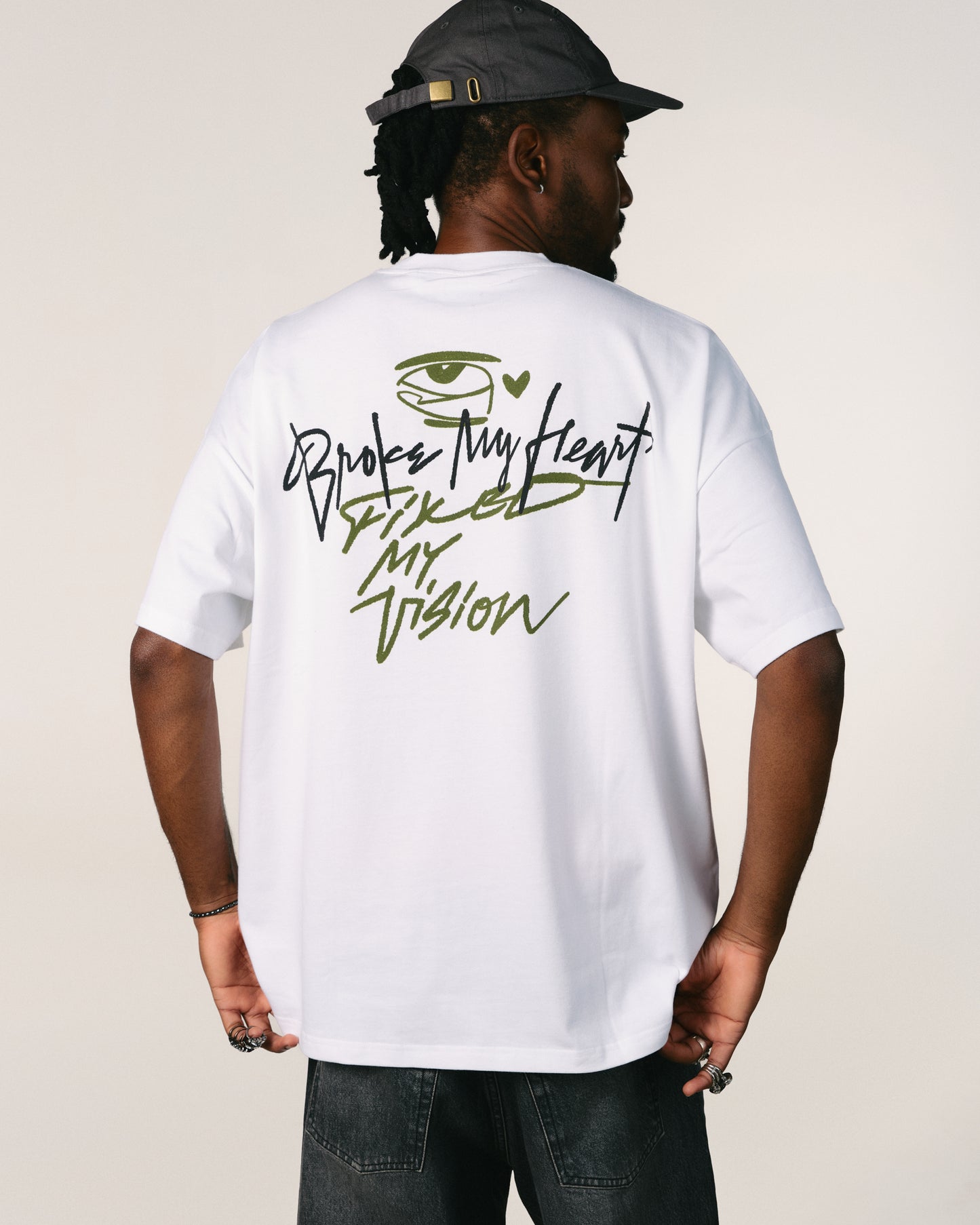 Person wearing a white hoppers hip hop takeovers t-shirt with text broke my heart fixed my vision and an eye design on the back, standing against a plain background Tricou oversized unisex Broke My Heart alb hip hop takeovers hoppers