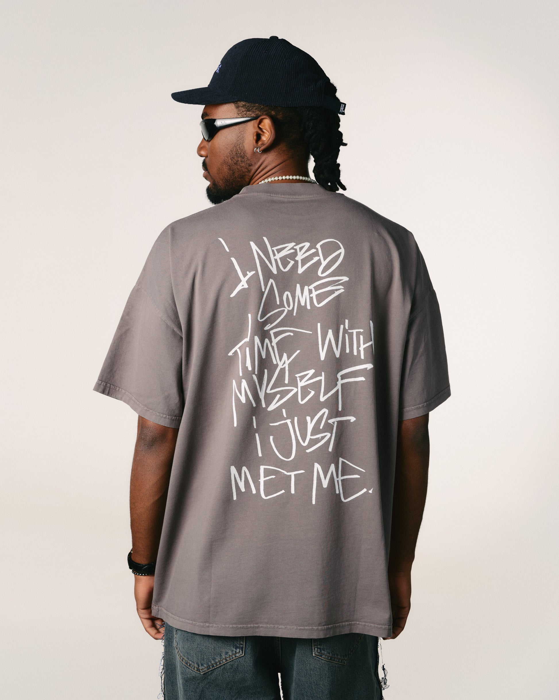 A person with long hair, wearing glasses, faces away, showcasing a Hoppers Hip Hop Takeovers T-shirt with graffiti text: "I need some time with myself I just met me." Casual, introspective vibe.