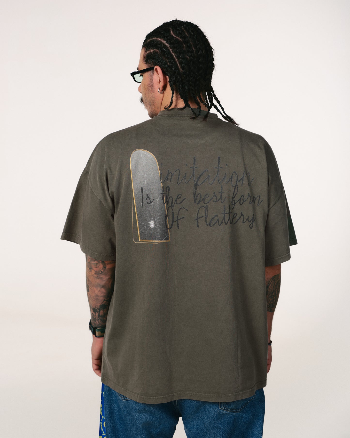Man with braided hair and tattoos wearing a gray T-shirt with the phrase "Imitation is the best form of flattery" written on the back. Casual tone.