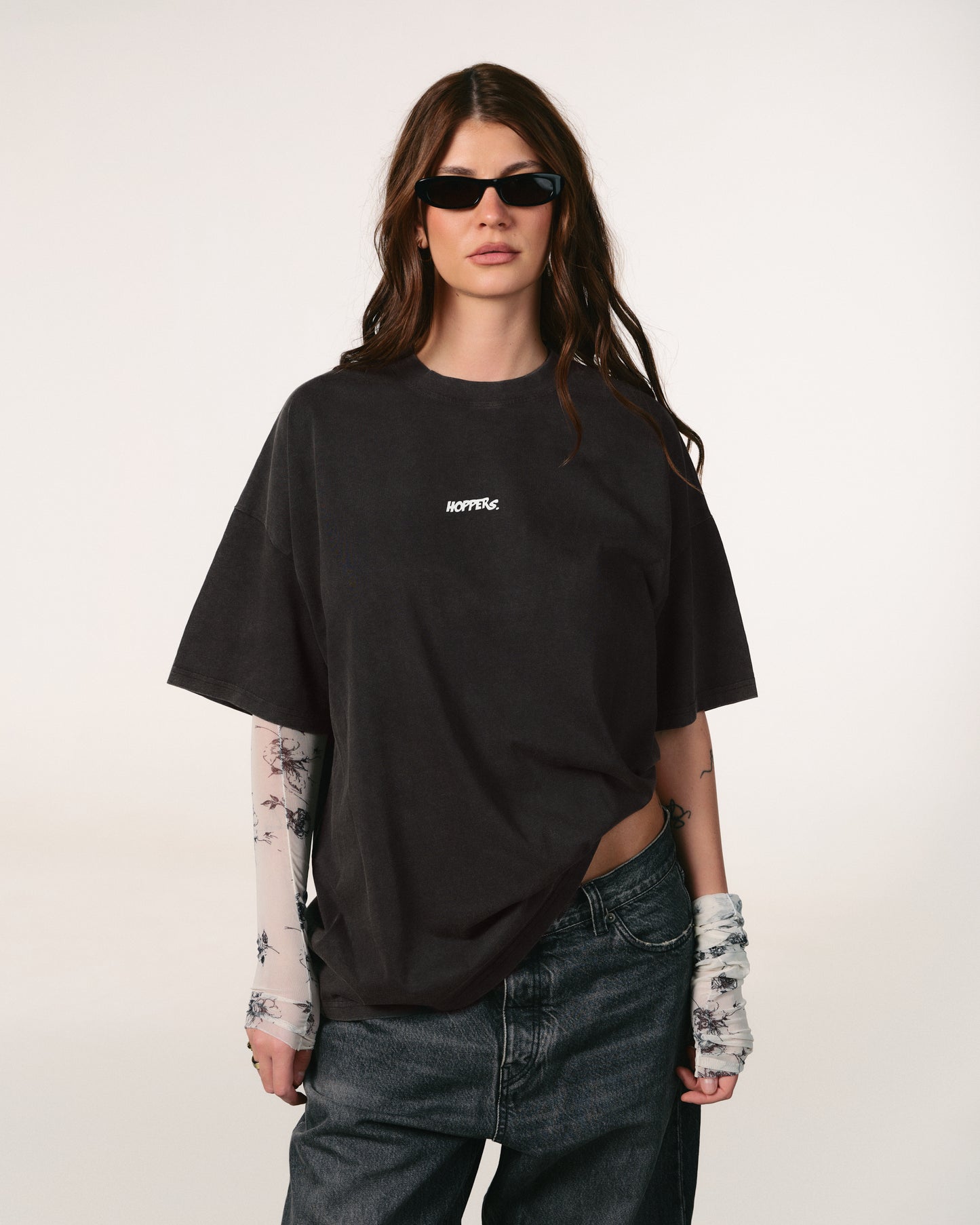 Person wearing a black oversized t-shirt with a brand logo, sunglasses, and tattoos on arms and hands against a white background. Tricou oversized unisex Hoppers Talking to Myself vintage grey, vedere frontala pe model, hip hop takeovers
