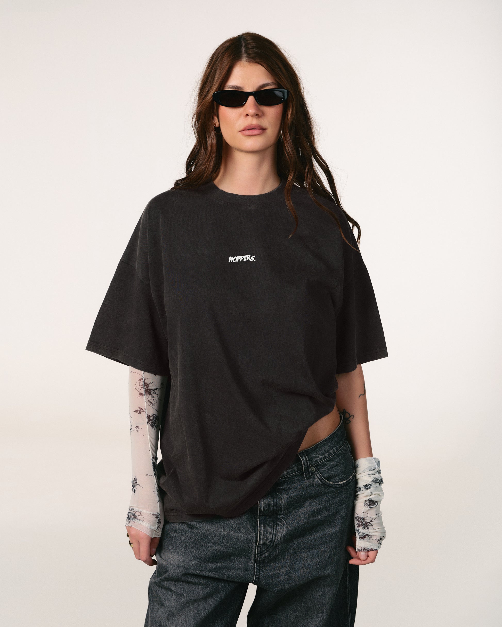 Person wearing a black oversized t-shirt with a brand logo, sunglasses, and tattoos on arms and hands against a white background. Tricou oversized unisex Hoppers Talking to Myself vintage grey, vedere frontala pe model, hip hop takeovers

