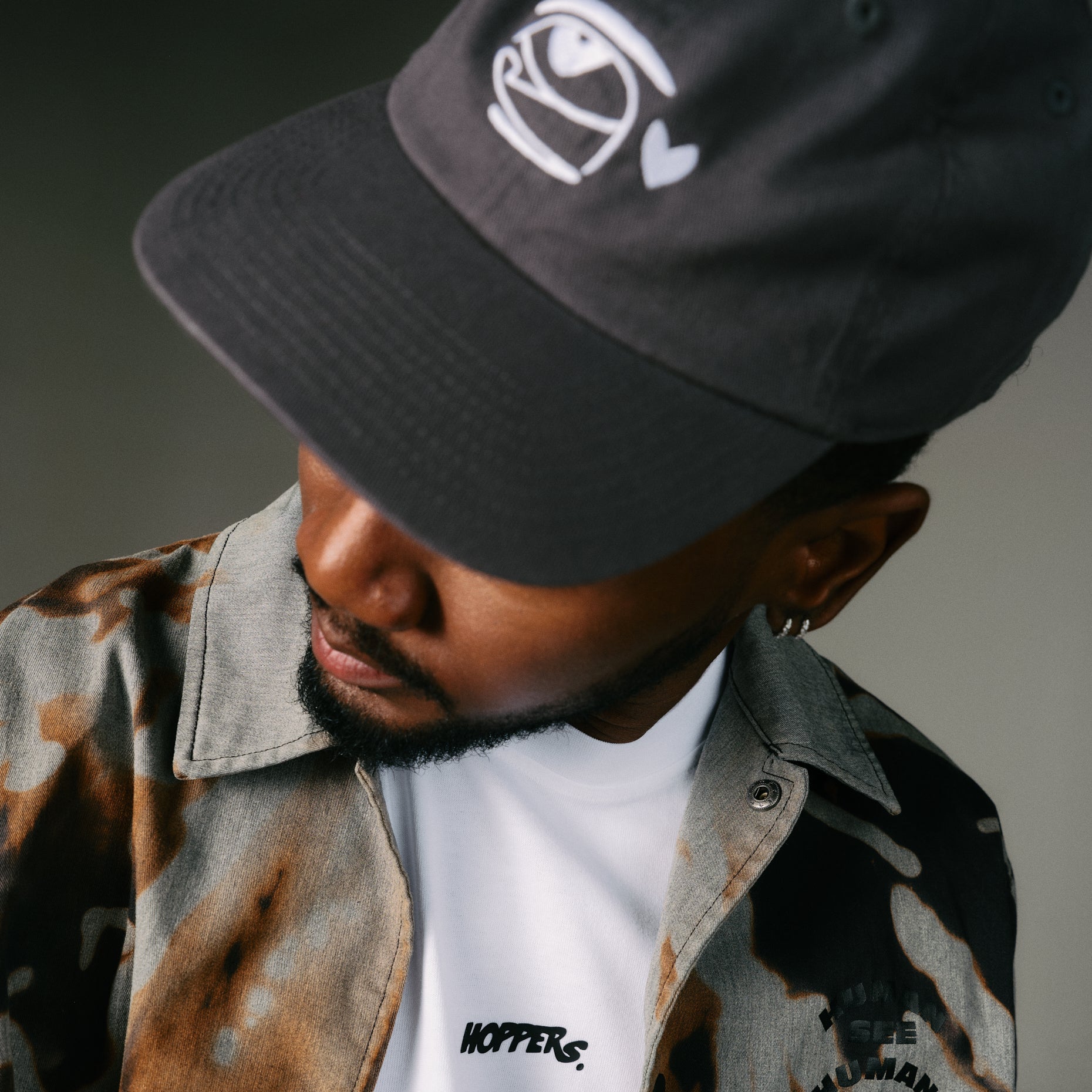 Man wearing a grey cap Hoppers hip hop takeovers with a logo and a patterned jacket over a white  hoppers shirt, 100% organic cotton.