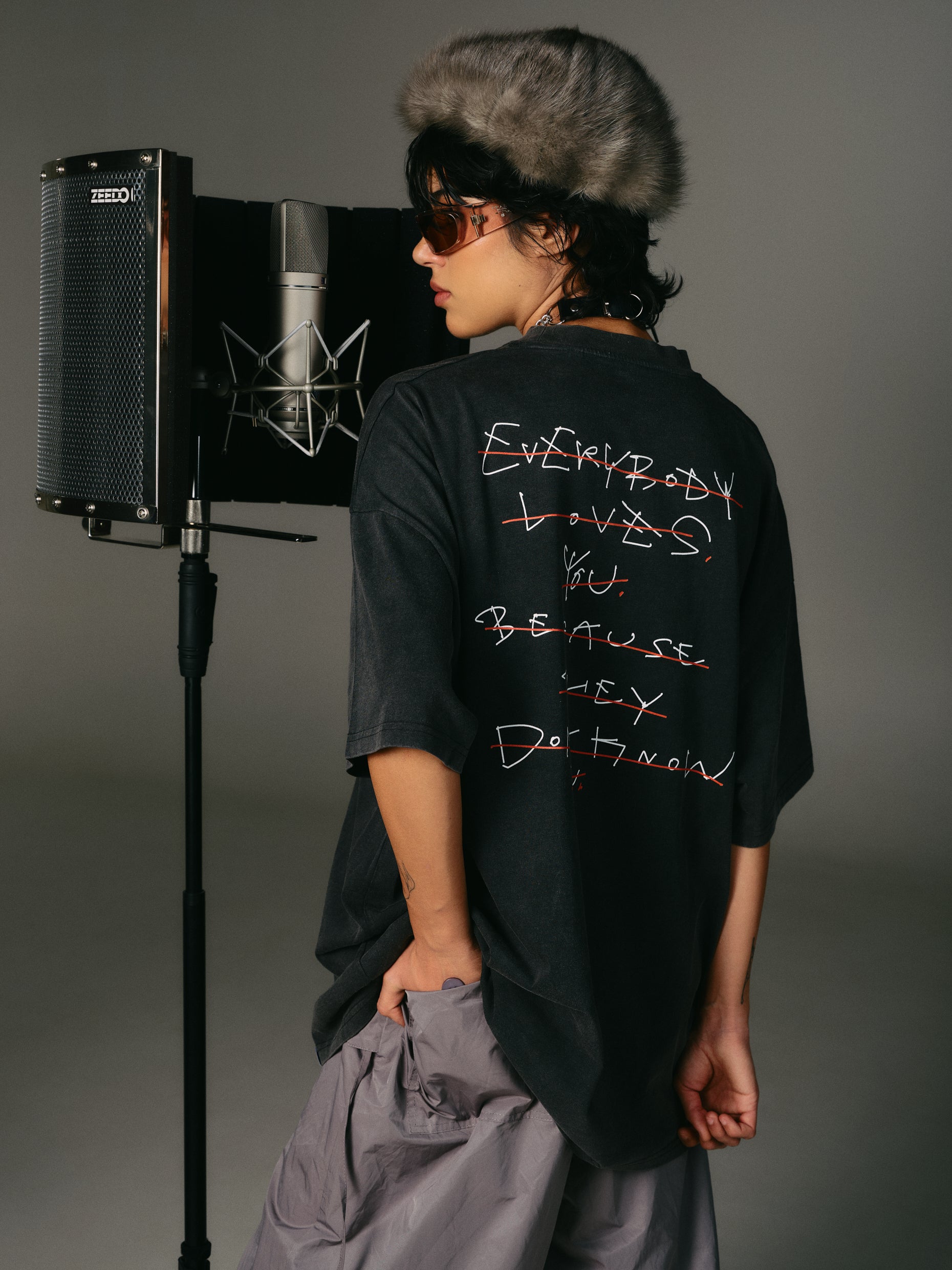 Person wearing a black hip hop takeovers hoppers t-shirt with text, unisex, standing in front of a microphone and speaker on a gray background